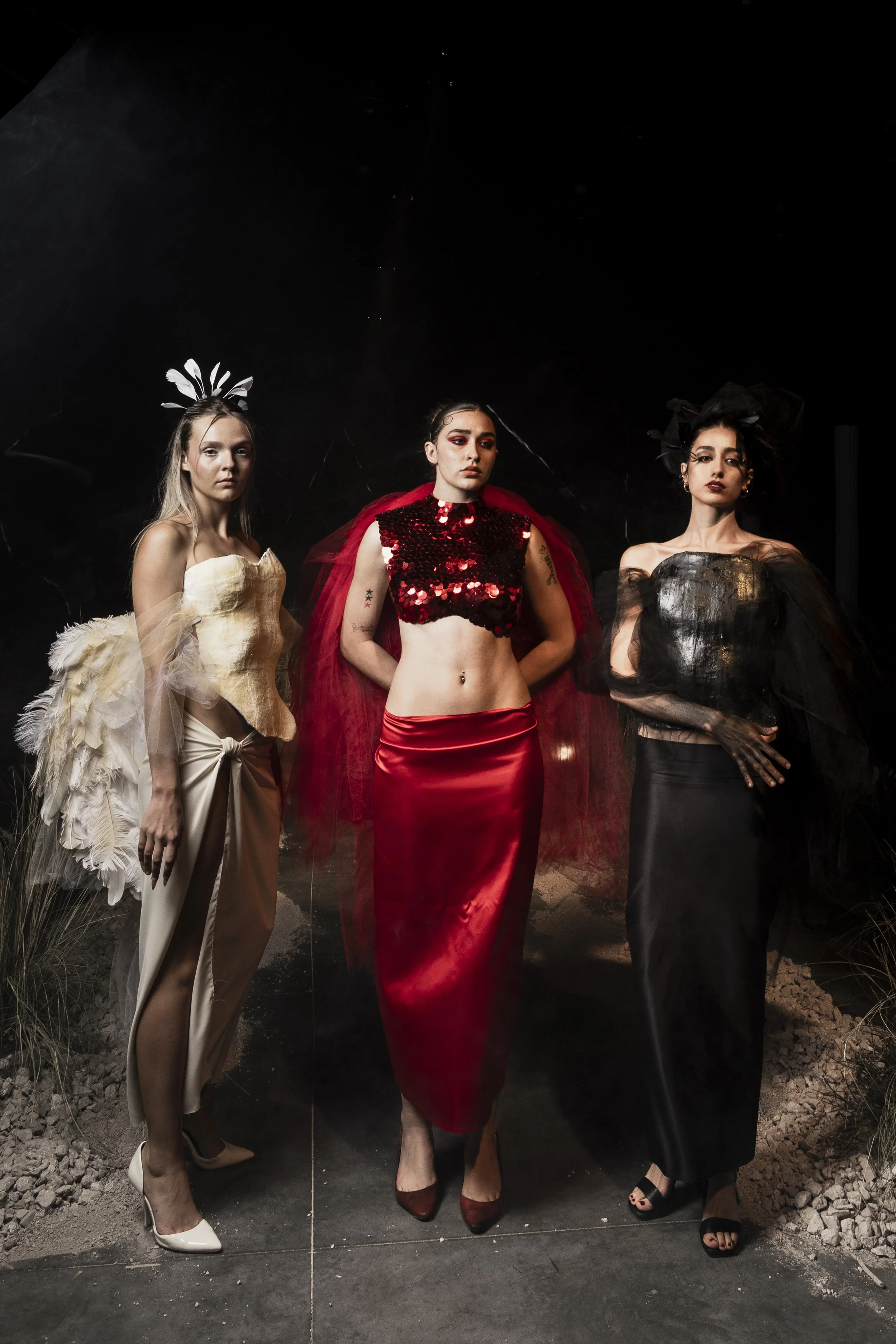 Three women in fashionable, elaborate outfits and accessories standing in a dark setting, with a ground of rocks and desert plants.
