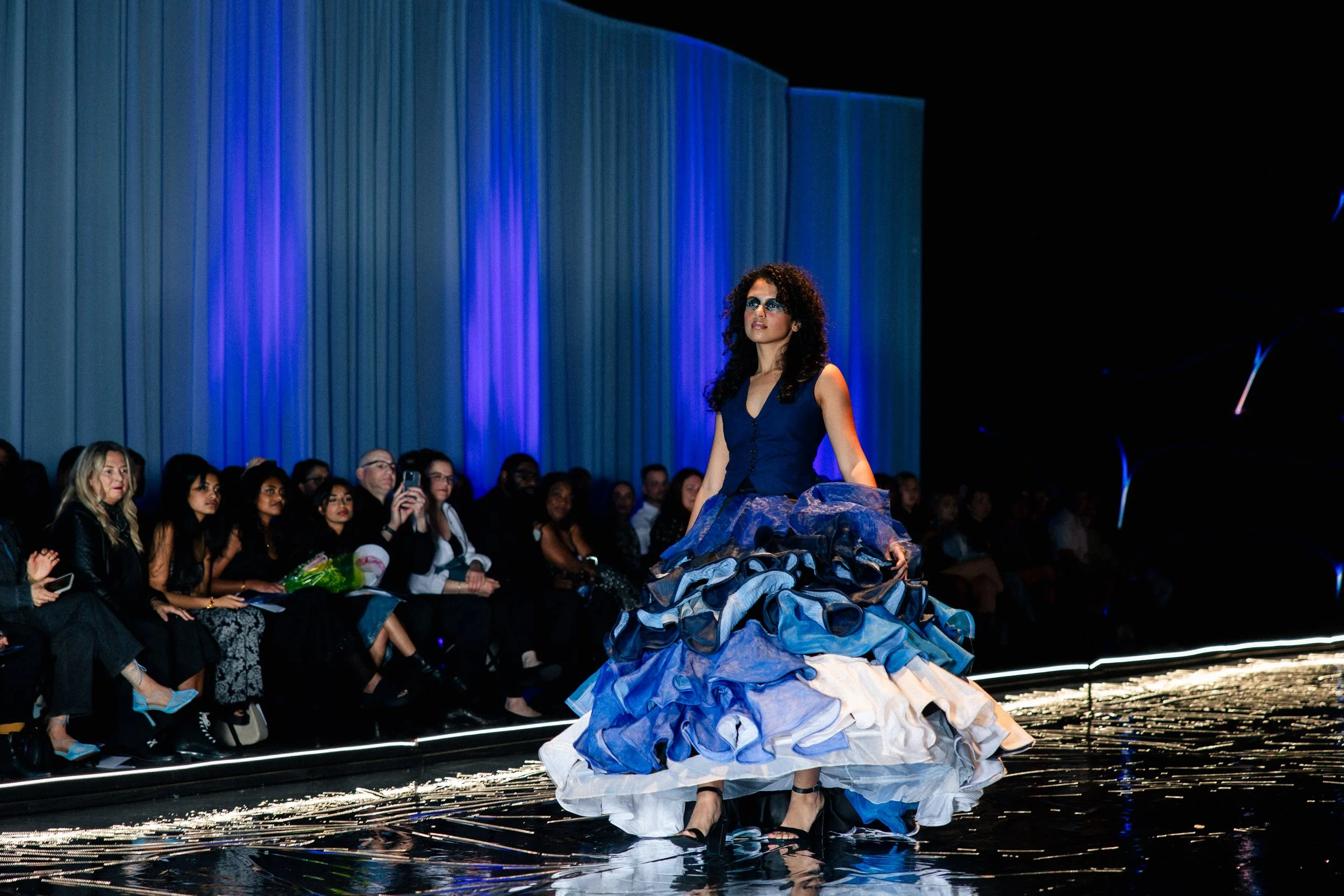 Model walking on runway in a blue and white layered dress, audience seated on either side, spotlight on the model, dark background with blue curtain.