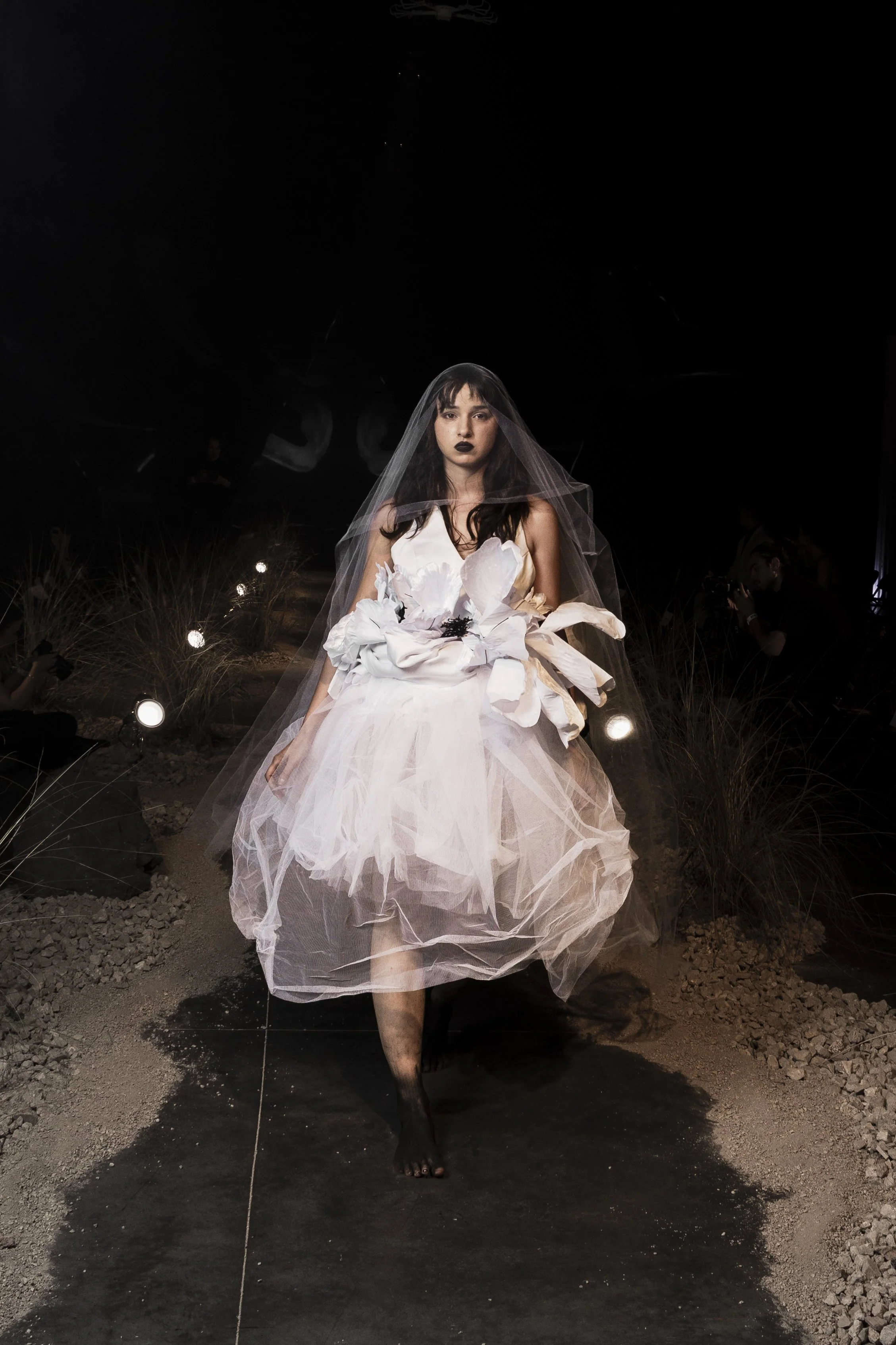A woman with dark hair, wearing black lipstick and a white dress with large floral details, is walking on a dark runway with lights and dry grass on each side, in a fashion show setting.