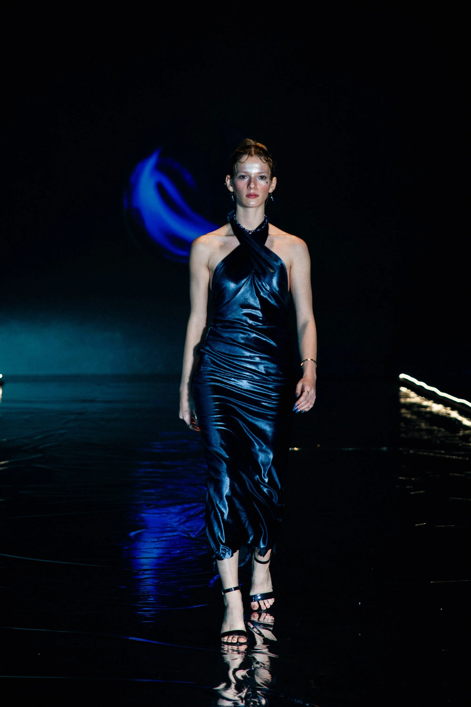 A model walking on a runway in a dark setting, wearing a navy blue satin gown with a halter neck and high heel sandals.