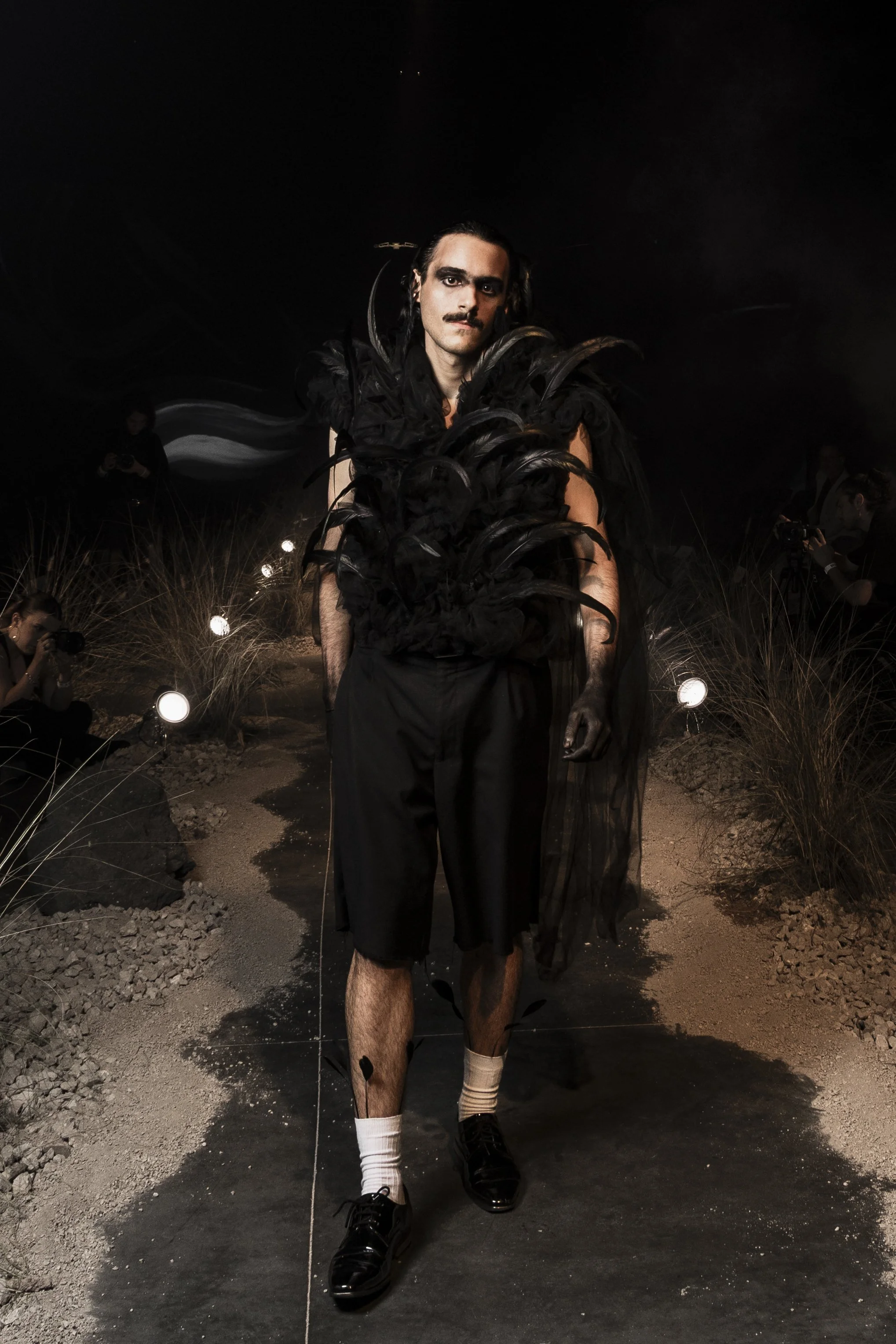 A person walking outdoors at night with dramatic makeup, black feathered shoulder accessories, dark clothing, white socks, and black shoes. They have dark hair and a mustache, surrounded by photographers and spotlights on a sandy path with grass.