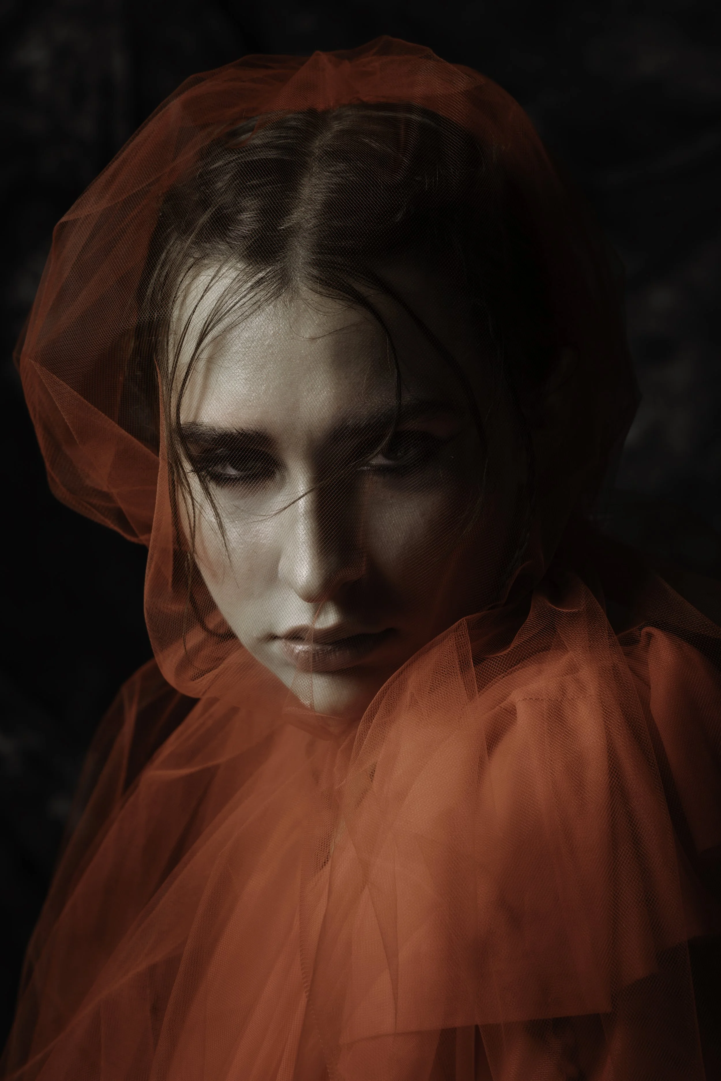 A woman with intense eye makeup and wet hair, wearing a red sheer fabric over her head and shoulders, against a dark background.