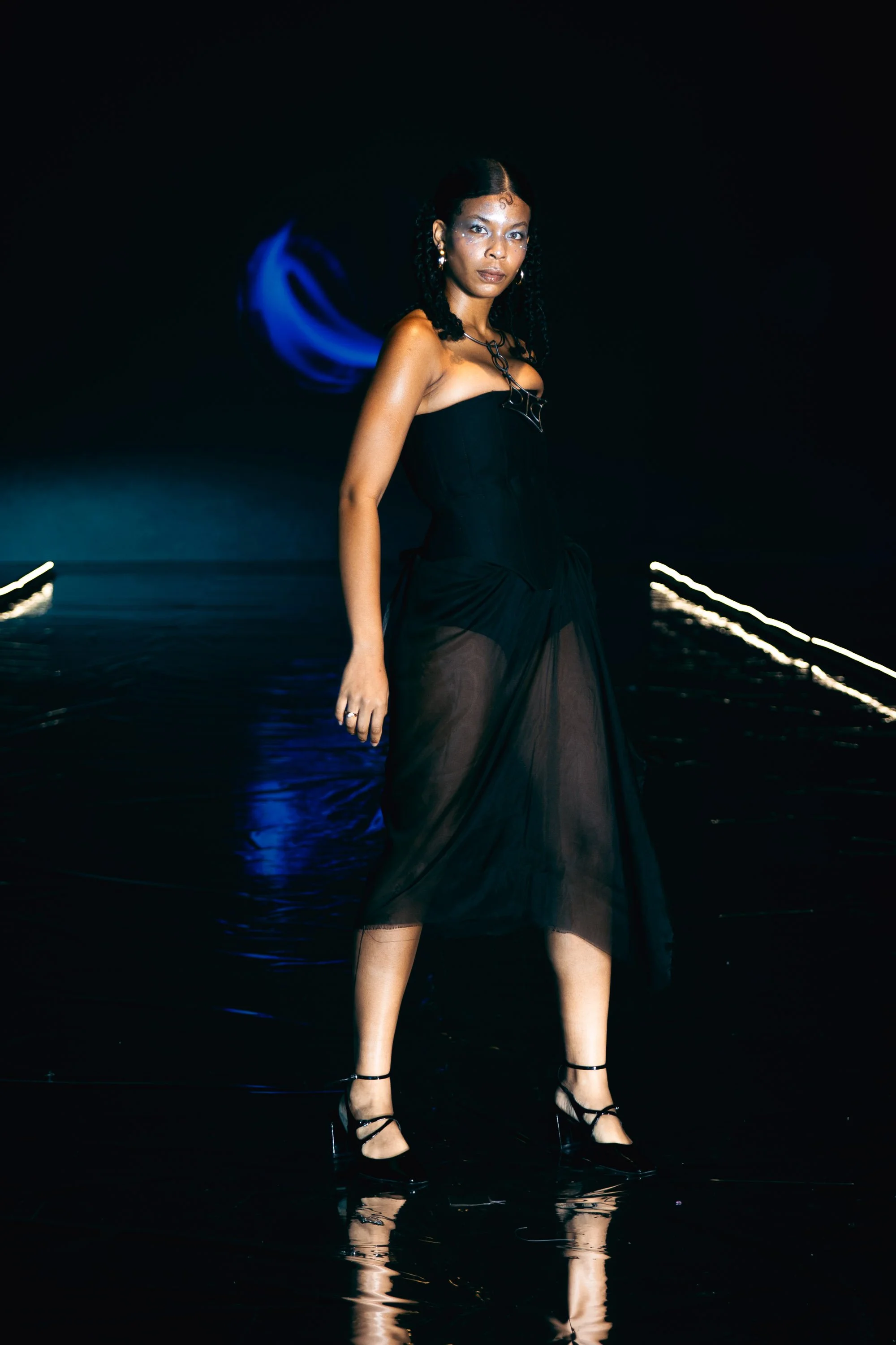 A woman walking on a dark runway, dressed in a black strapless dress with a sheer overlay, wearing black high heels, with a blue light in the background.
