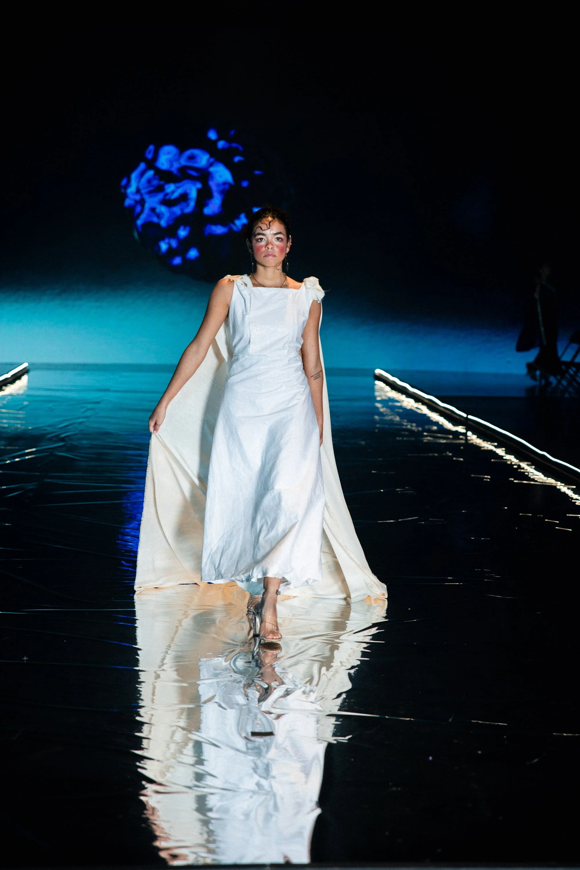 A woman in a white dress walking on a reflective runway with water. She is illuminated with blue lighting in the background, which features an abstract blue pattern projected on the wall.
