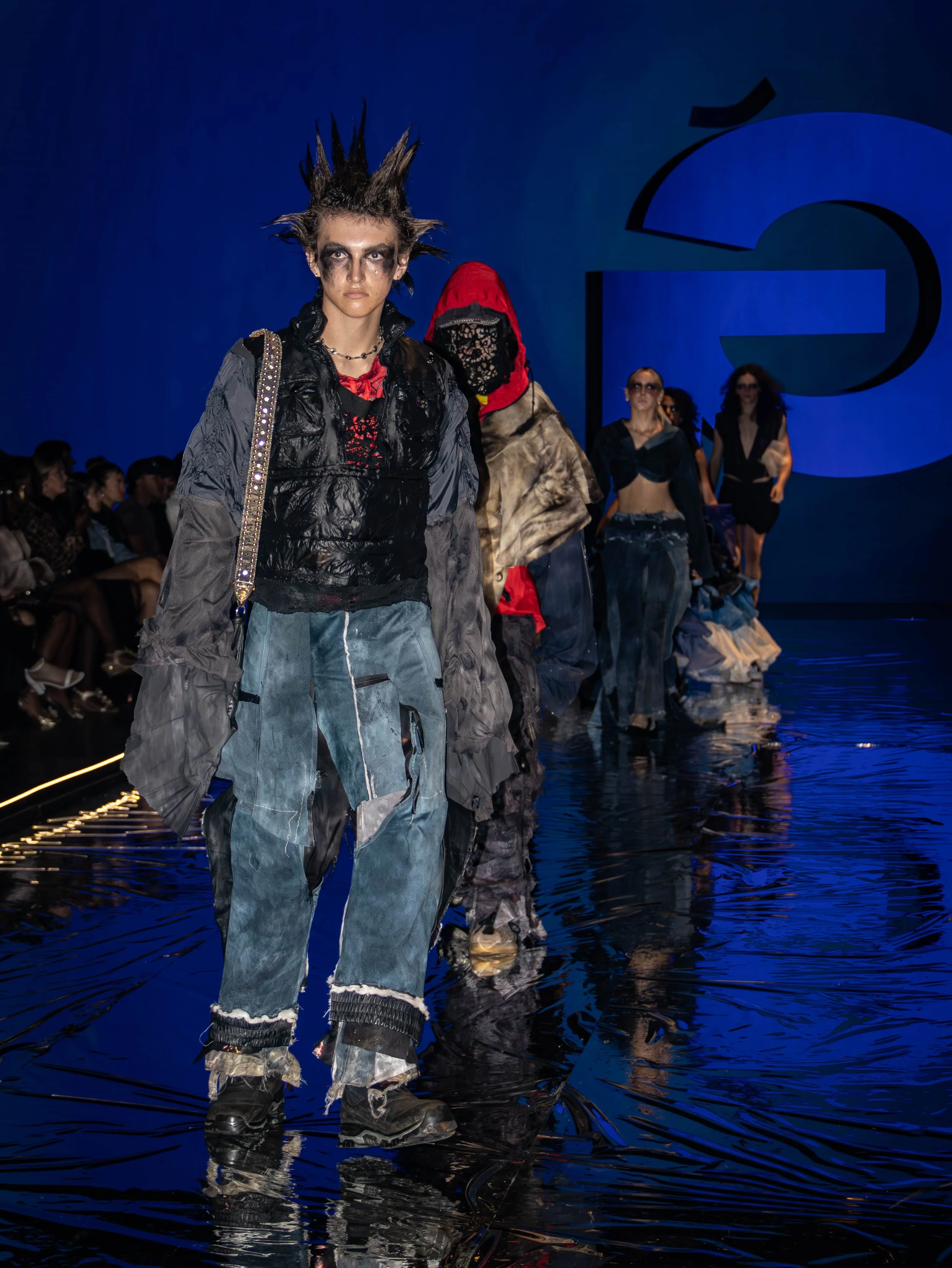 Models walk on a runway at a fashion show, wearing avant-garde outfits with dark, edgy makeup, and the background features a large blue logo on a dark backdrop.