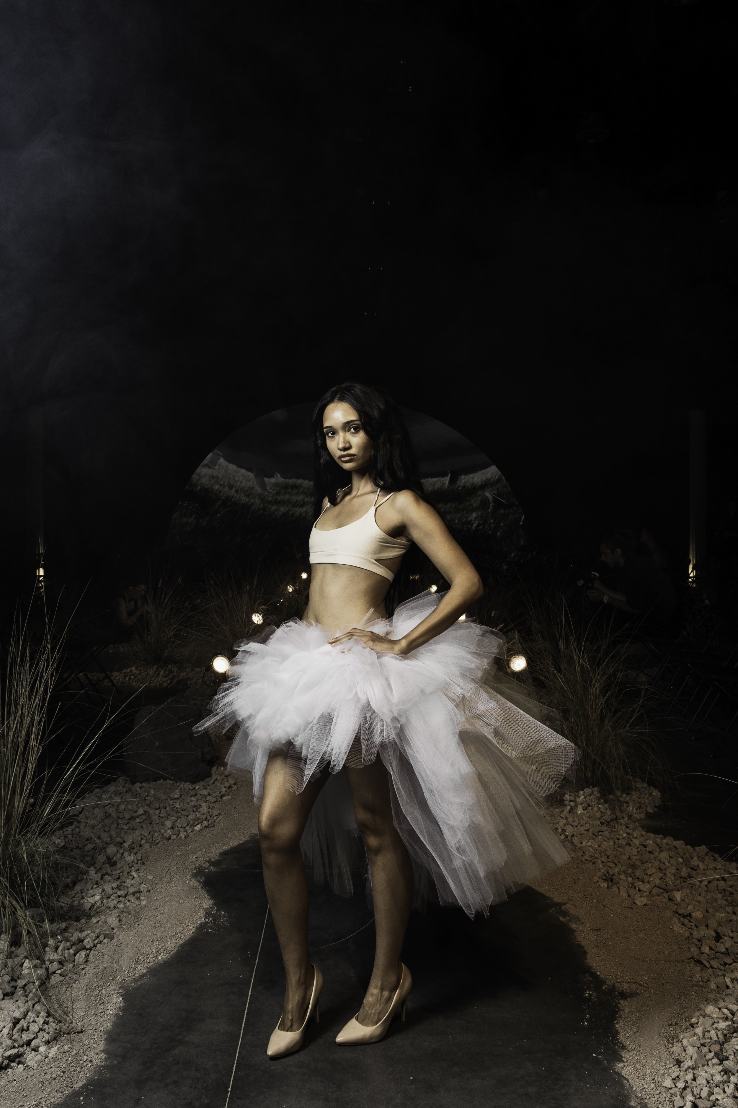 A woman in a white ballet costume with a tulle skirt is standing in a dark outdoor setting at night, with decorative lighting and a pathway surrounded by rocks and plants.