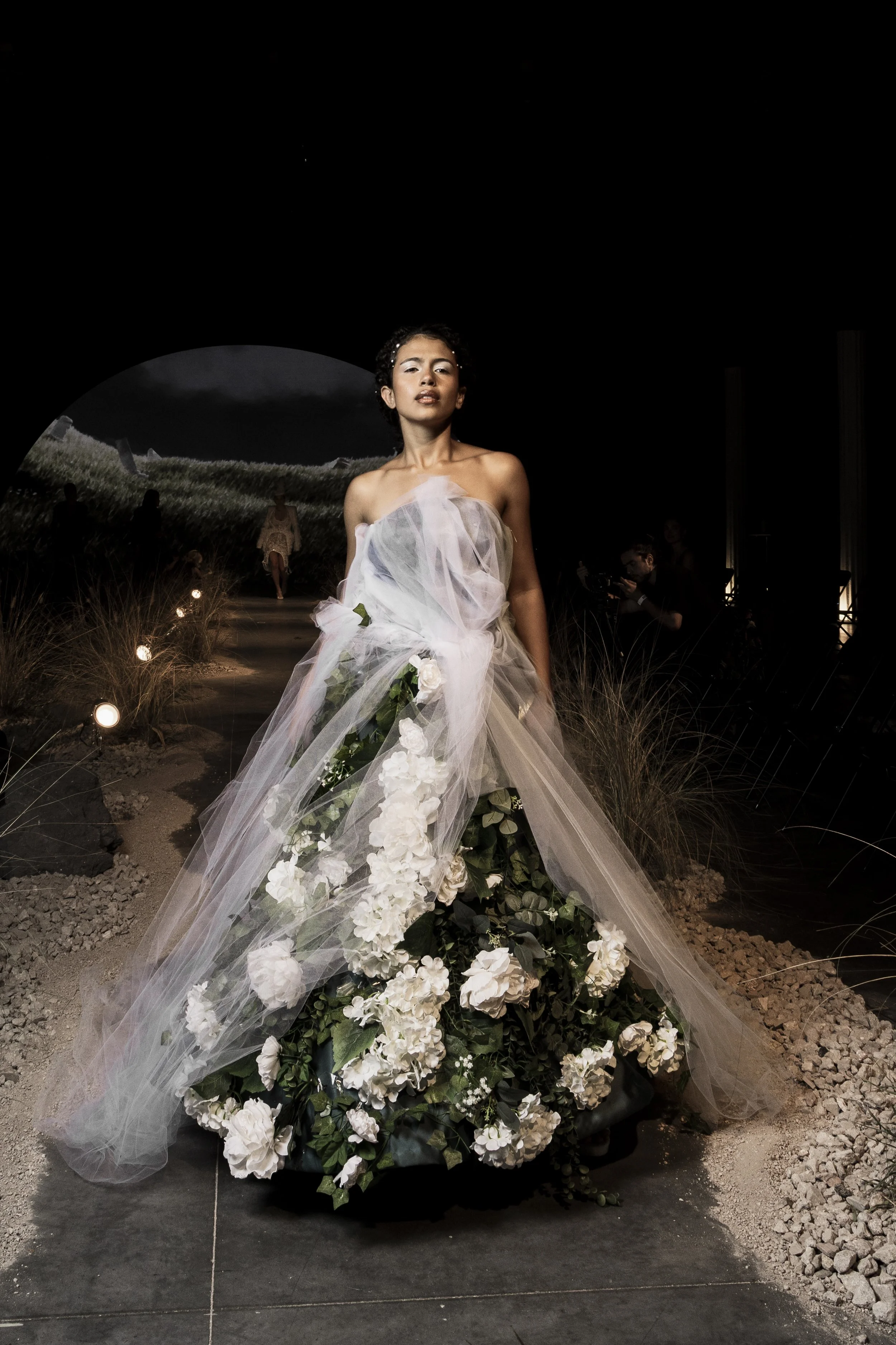 A fashion model on a runway wearing a strapless gown made of sheer, flowing fabric with white flowers and green leaves, against a dark background with a landscape scene projected behind her, accompanied by a photographer capturing the moment.