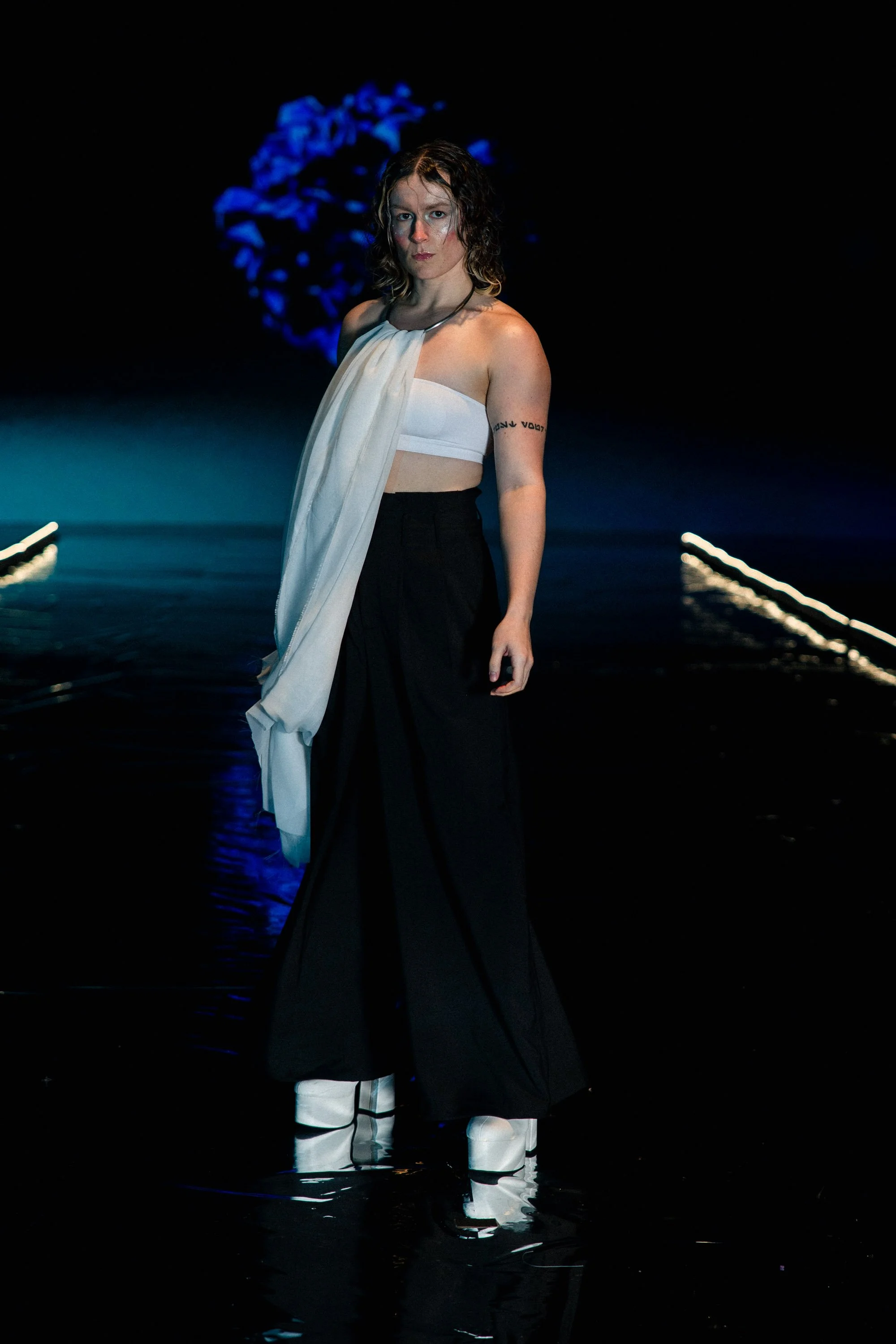 Model walking on a runway during a fashion show, wearing a white strapless top with an attached draped fabric, black wide-leg pants, and white platform shoes, with a dark background and blue lighting.