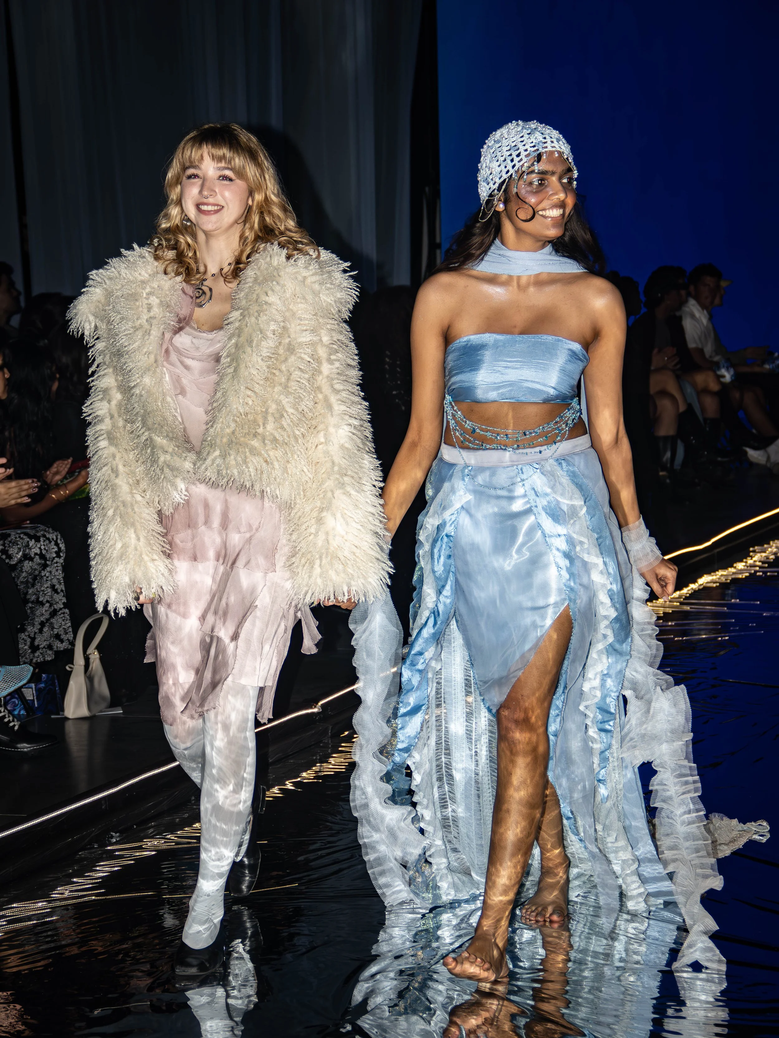 Two women walk hand-in-hand on a runway during a fashion show, wearing elaborate outfits against a dark backdrop with audience members watching.