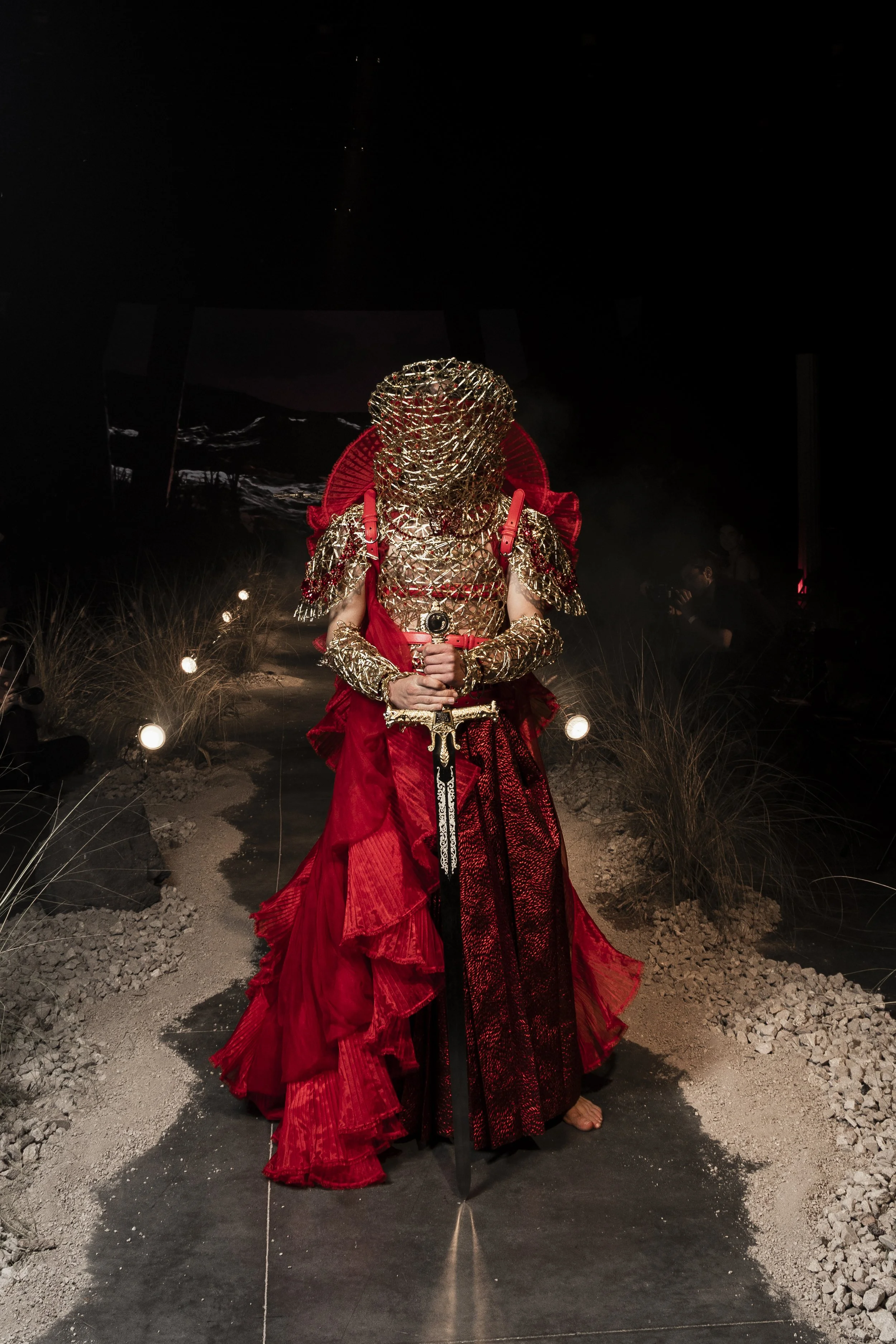 Person in elaborate costume with gold lattice headpiece and red gown, standing in a dark setting with spotlights and dry grass decor.