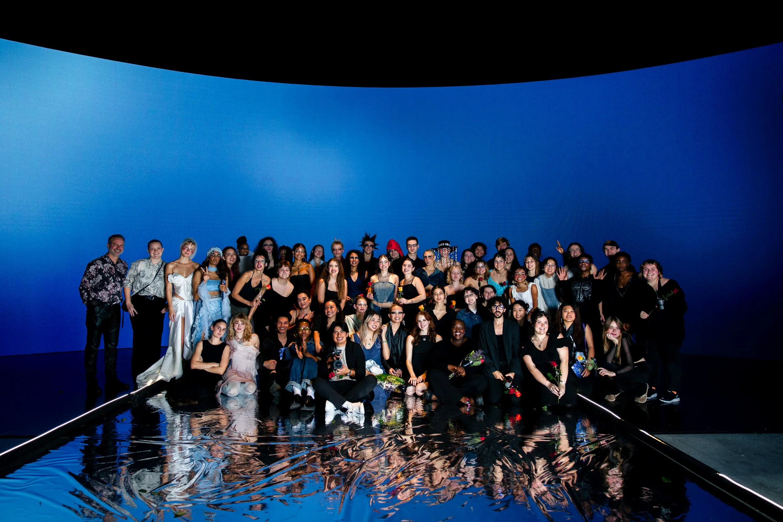 A large group of people posing together on a reflective floor in front of a deep blue curved wall, with some holding flowers and others making peace signs.