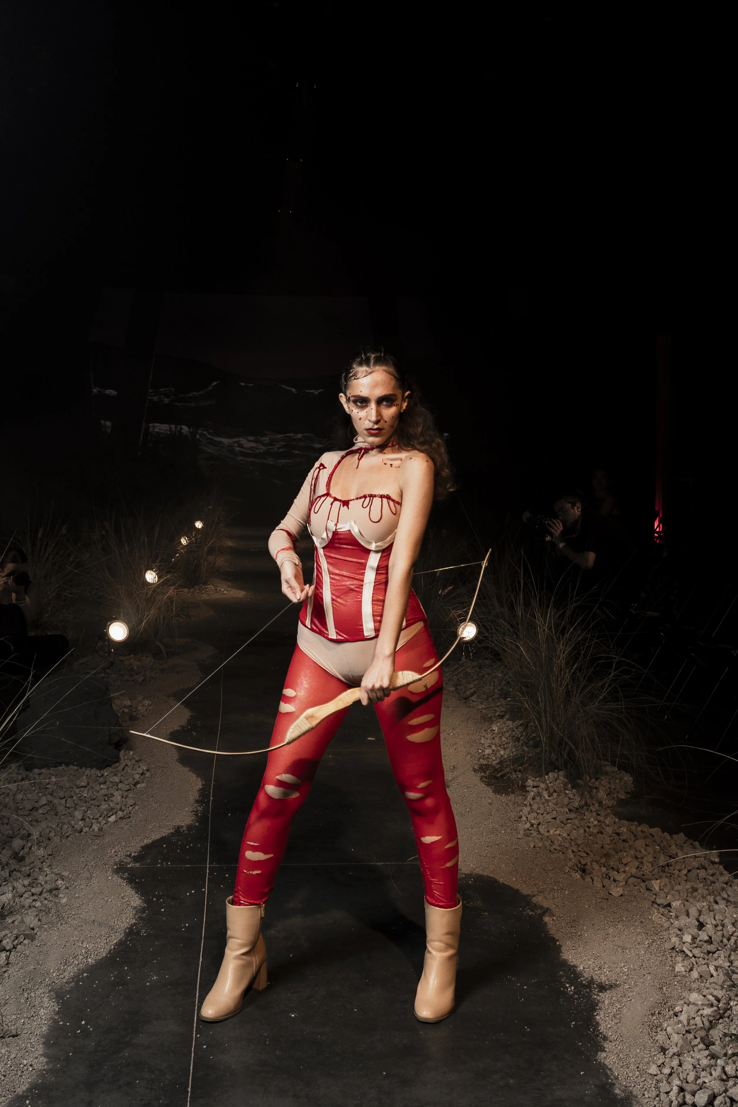 A woman with makeup and a red latex outfit holding a bow, standing outdoors at night on a dark path with spotlights and plants.