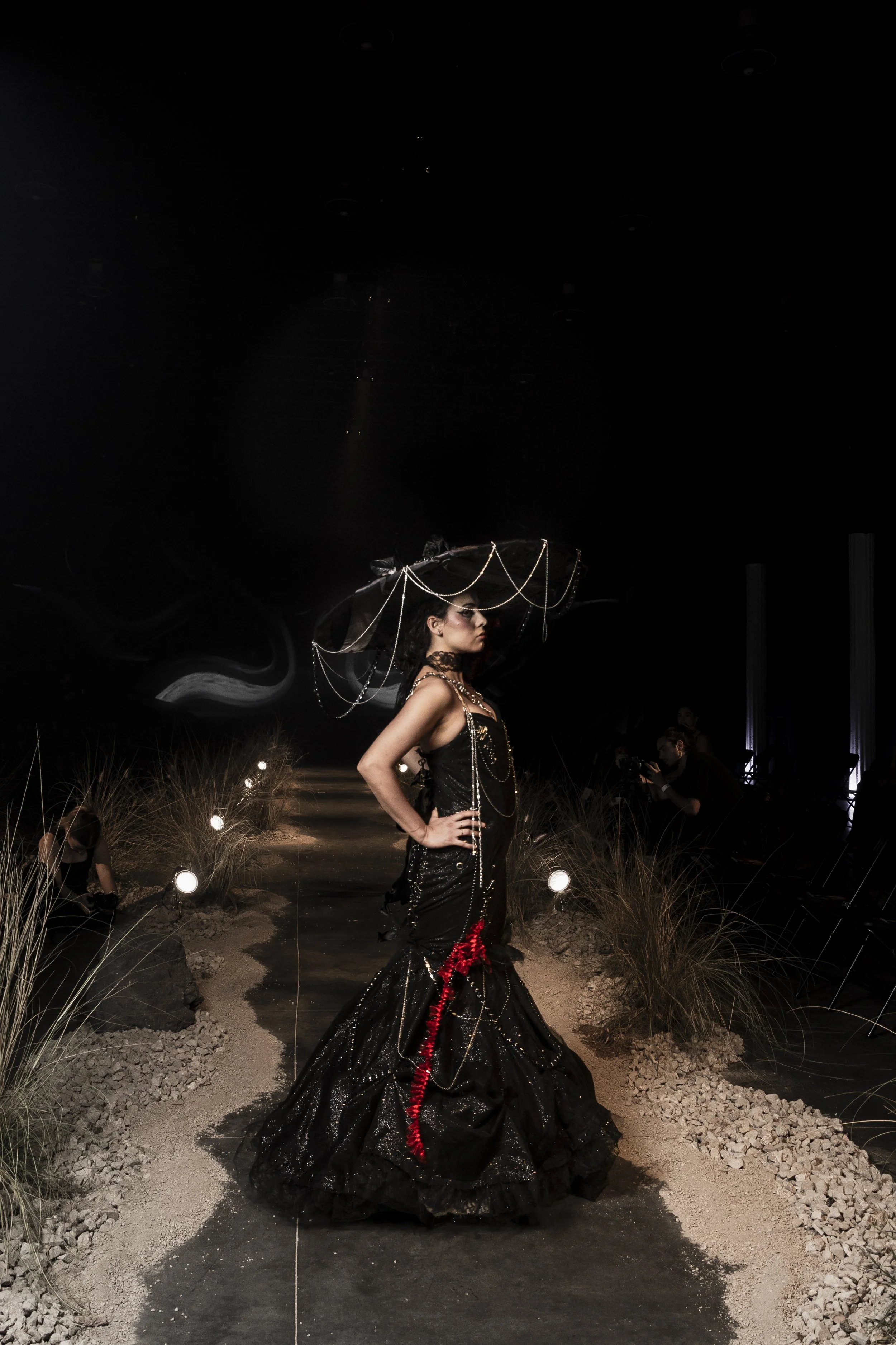 A woman walking on a runway at night in a black gown with red accents and jewelry, wearing a large black hat with decorative chains, surrounded by grass and rocks, with photographers taking pictures.