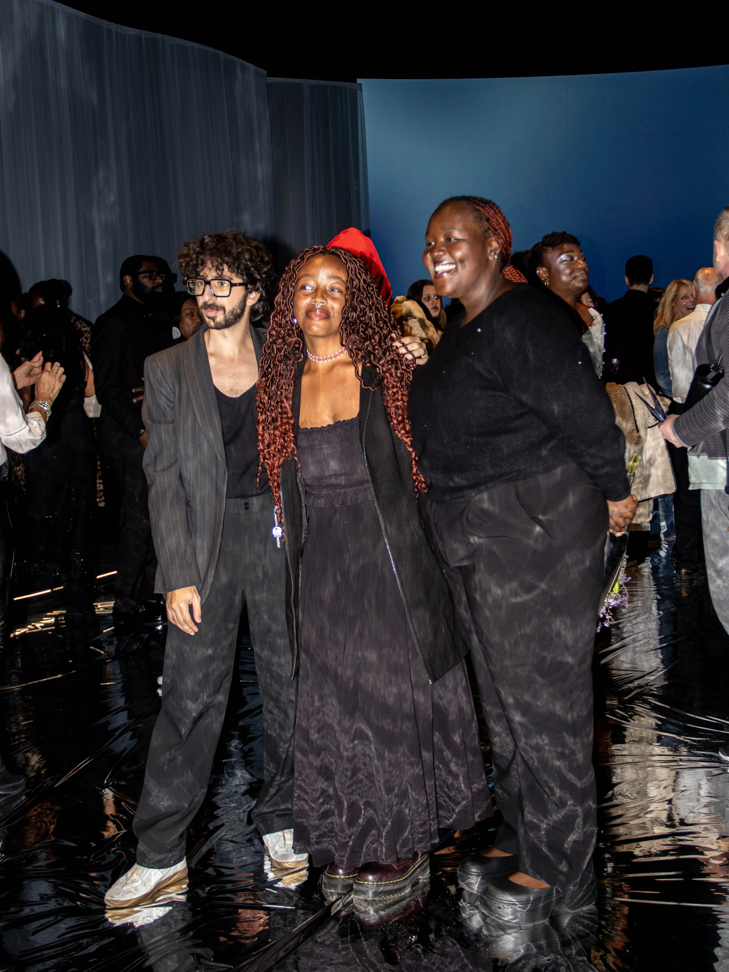 Group of diverse people at an art or fashion event, standing on a shiny black floor with a dark, abstract background.