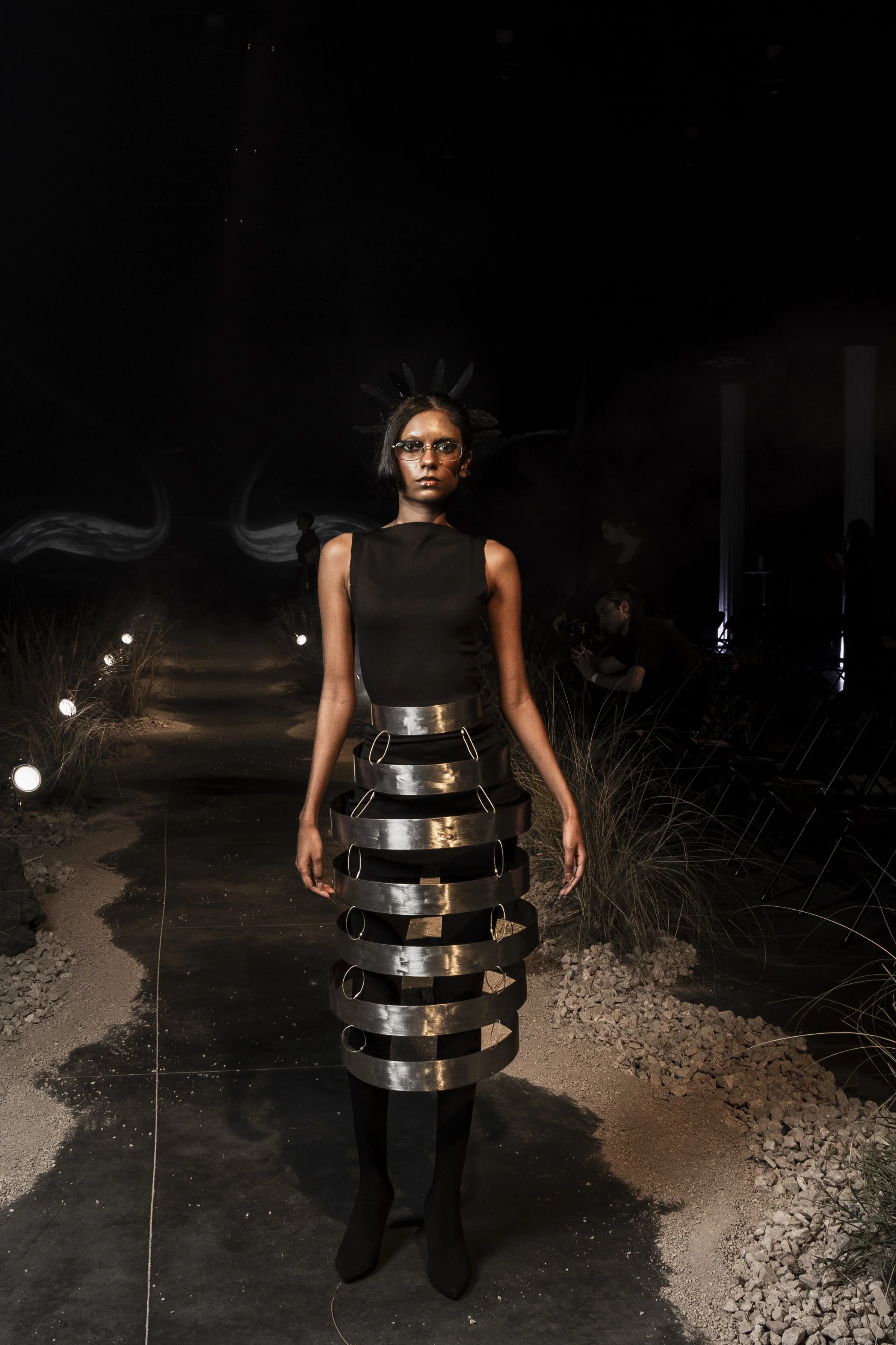 A female model walking on a runway during a fashion show, wearing a black sleeveless top and a unique metallic skirt with circular cutouts, set against a dark background with subtle lighting.