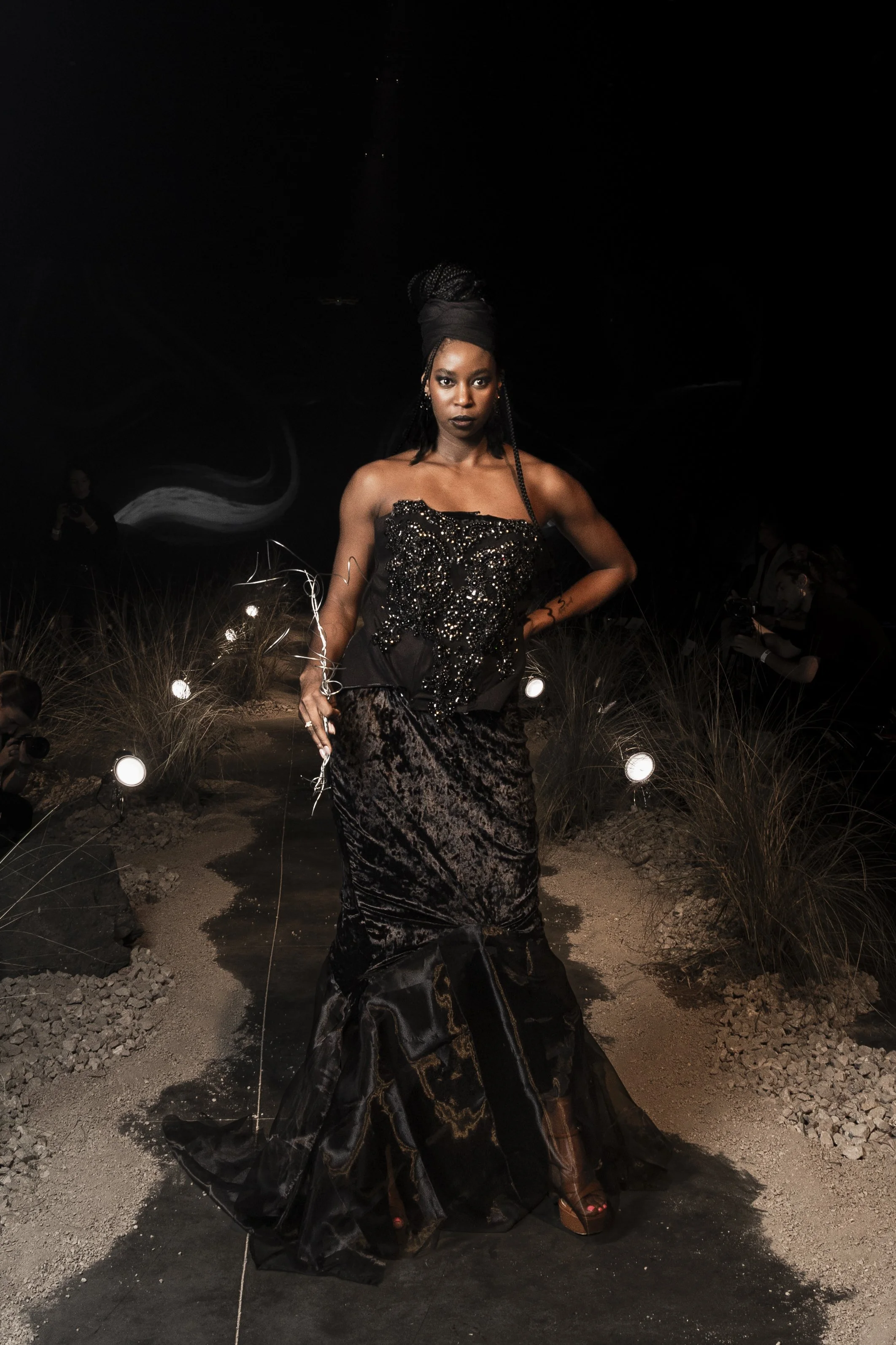 A woman with dark skin and long braided hair, wearing a black strapless gown with intricate beading and a full velvet skirt, stands on a dark outdoor pathway illuminated by small lights, with a black background and some other people faintly visible i