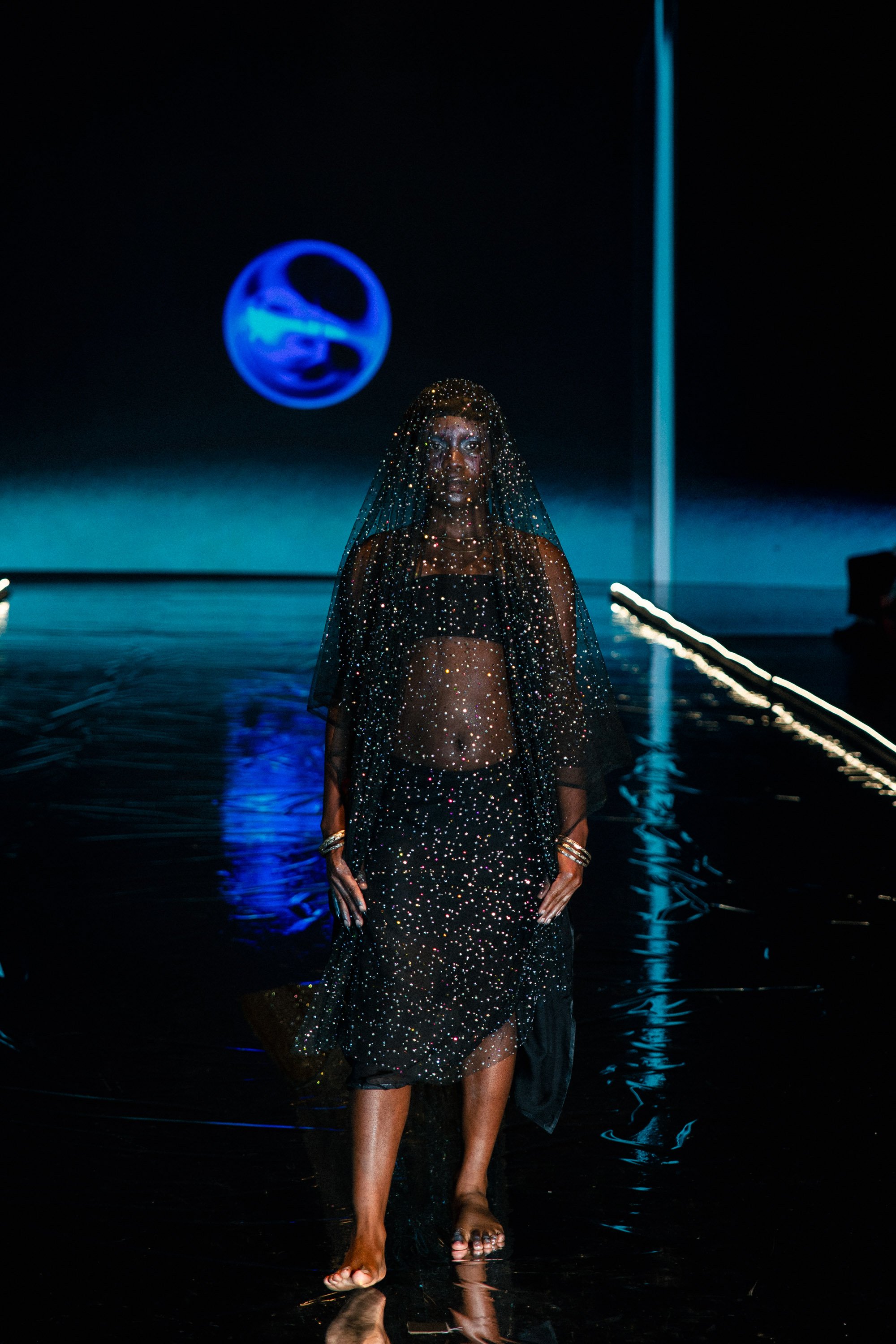A woman walking on a stage in a black, sparkling outfit with a veil, backdrop featuring blue and black lighting, and a spherical holographic display.