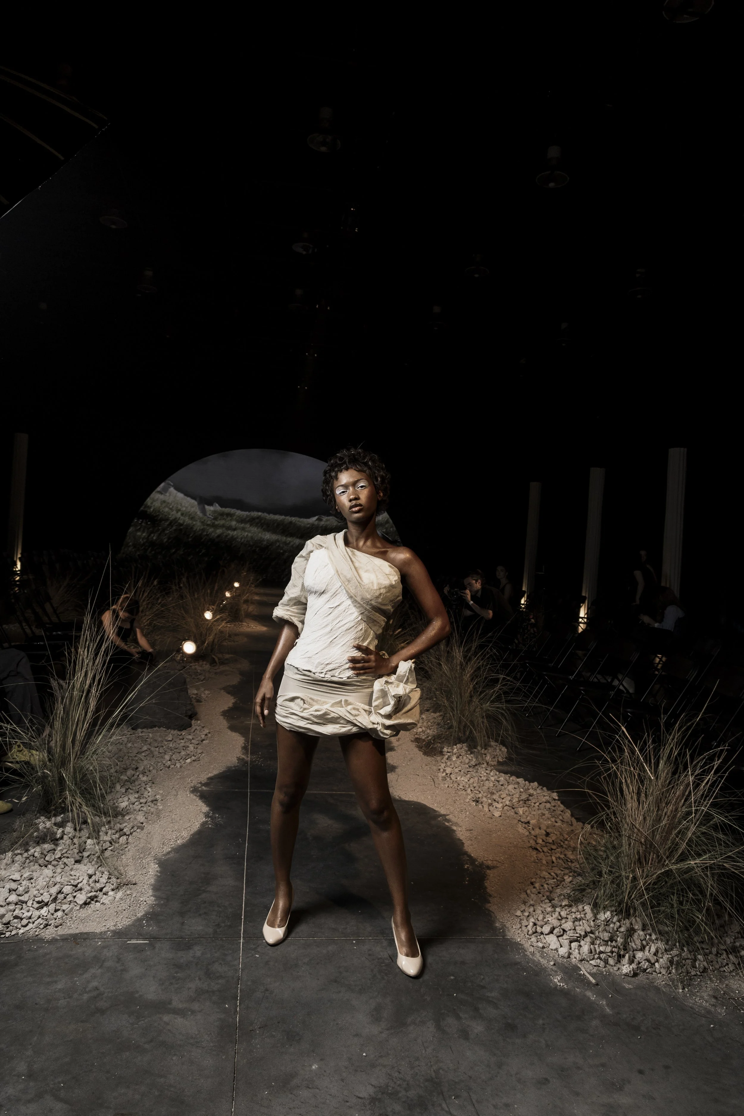 Fashion model on runway wearing beige textured dress, standing in dark indoor setting with decorative grass and landscape background.