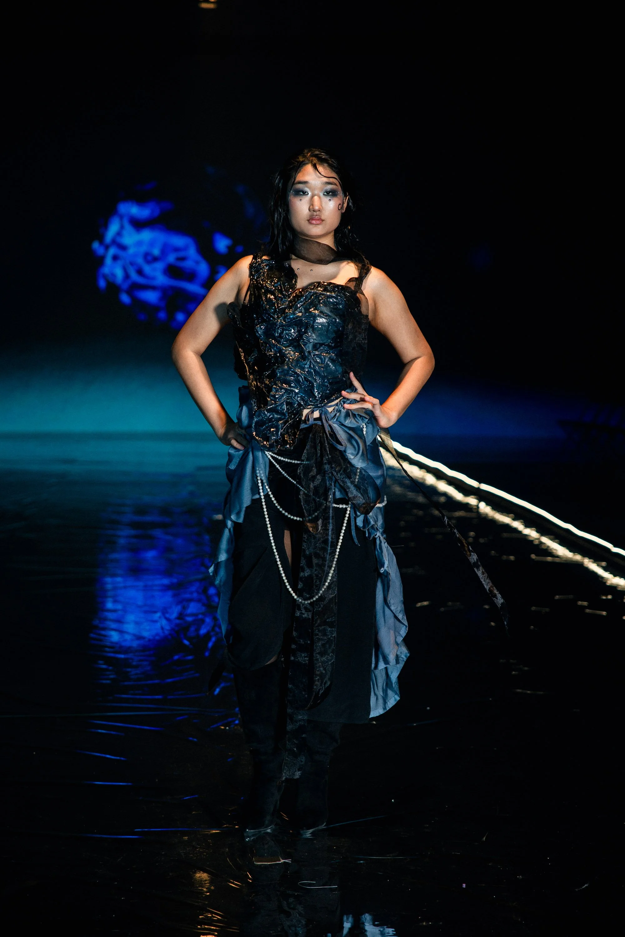 A model walking down a runway during a fashion show, wearing a dark, avant-garde dress with textured fabric, pearls, and layered materials, with a dark background and blue lighting.