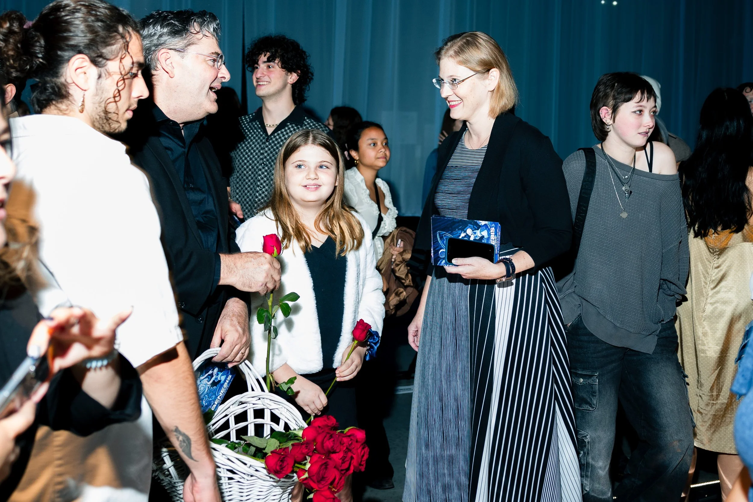 A group of people, including a man, a woman, and a young girl holding red roses, are engaged in conversation at an indoor event with a blue curtain in the background. Others are nearby, some holding bags or wearing casual clothing.