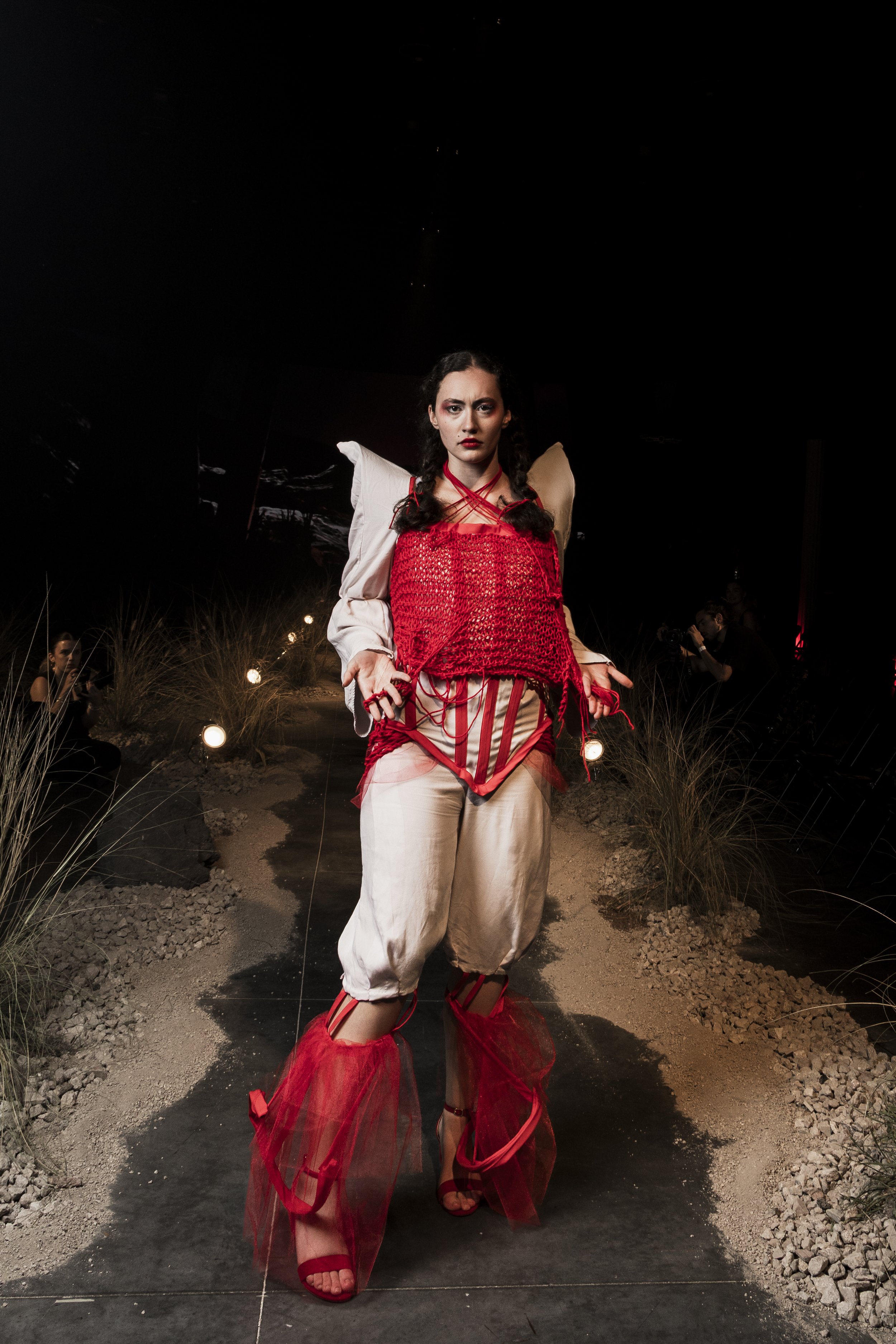 A woman walking down a dark pathway decorated with small lights, wearing a red and beige fashion-forward outfit with layered tulle and woven fabric, and red high-heeled shoes, in an outdoor setting at night.