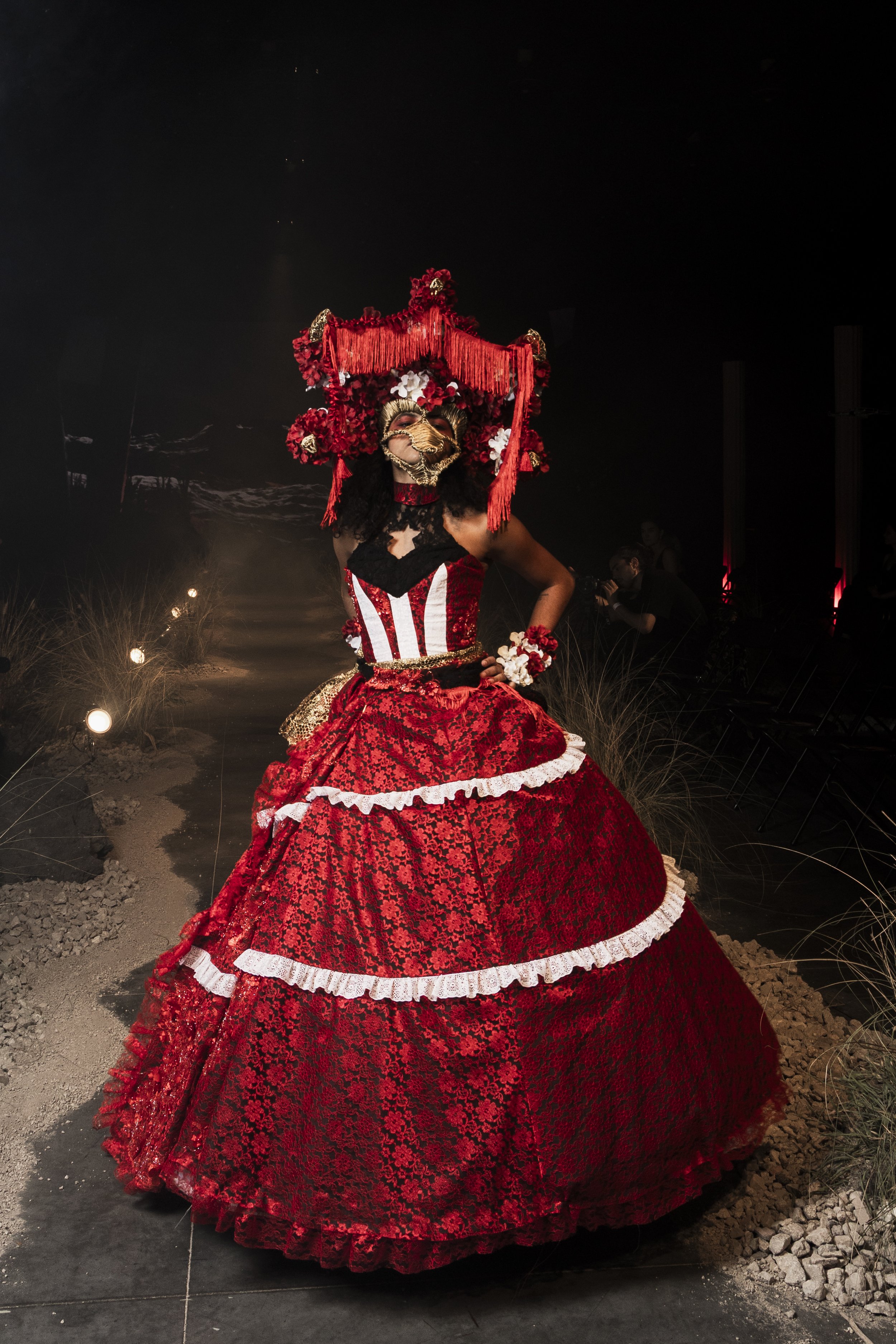 A person wearing a detailed red dress with black and white accents, and an elaborate headpiece decorated with flowers, tassels, and a mask with a beak, standing on a dark stage with minimal lighting.