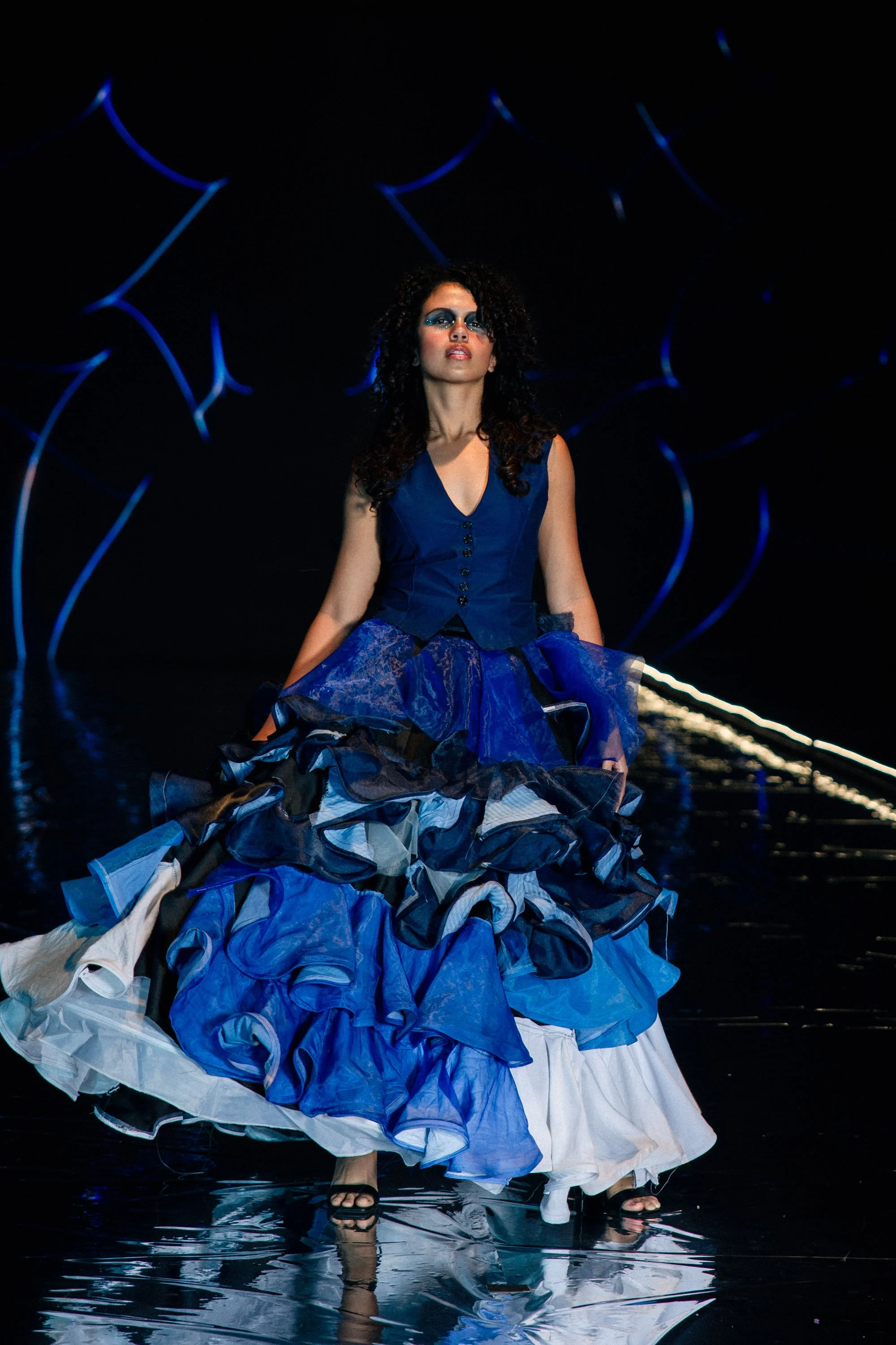 Fashion model wearing a blue and white ruffled dress on runway with dark background and abstract blue light design.