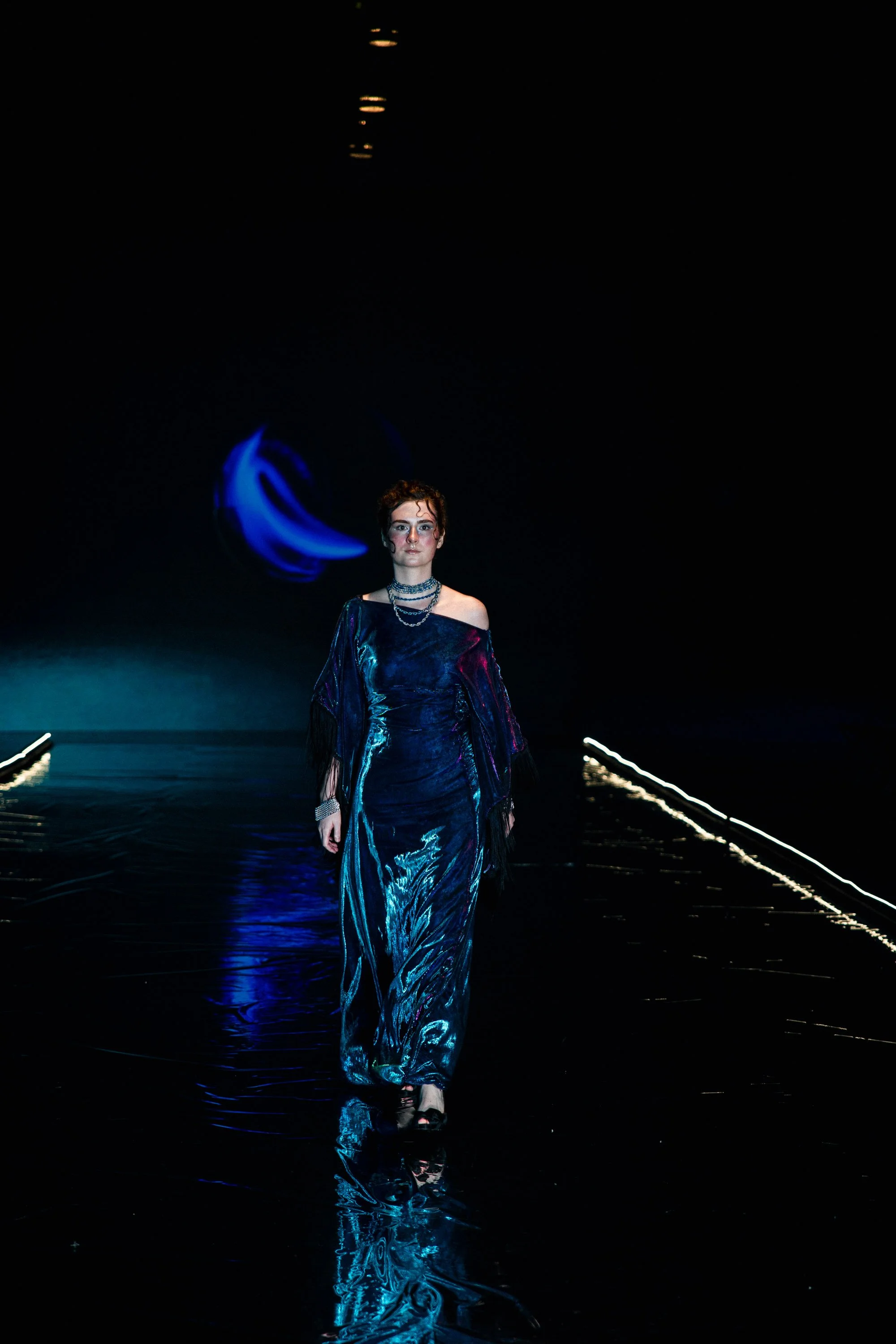 A model walking down a runway wearing a shiny, flowing, dark blue dress with metallic accents, accessorized with layered necklaces and bracelets, against a dark background with minimal lighting.