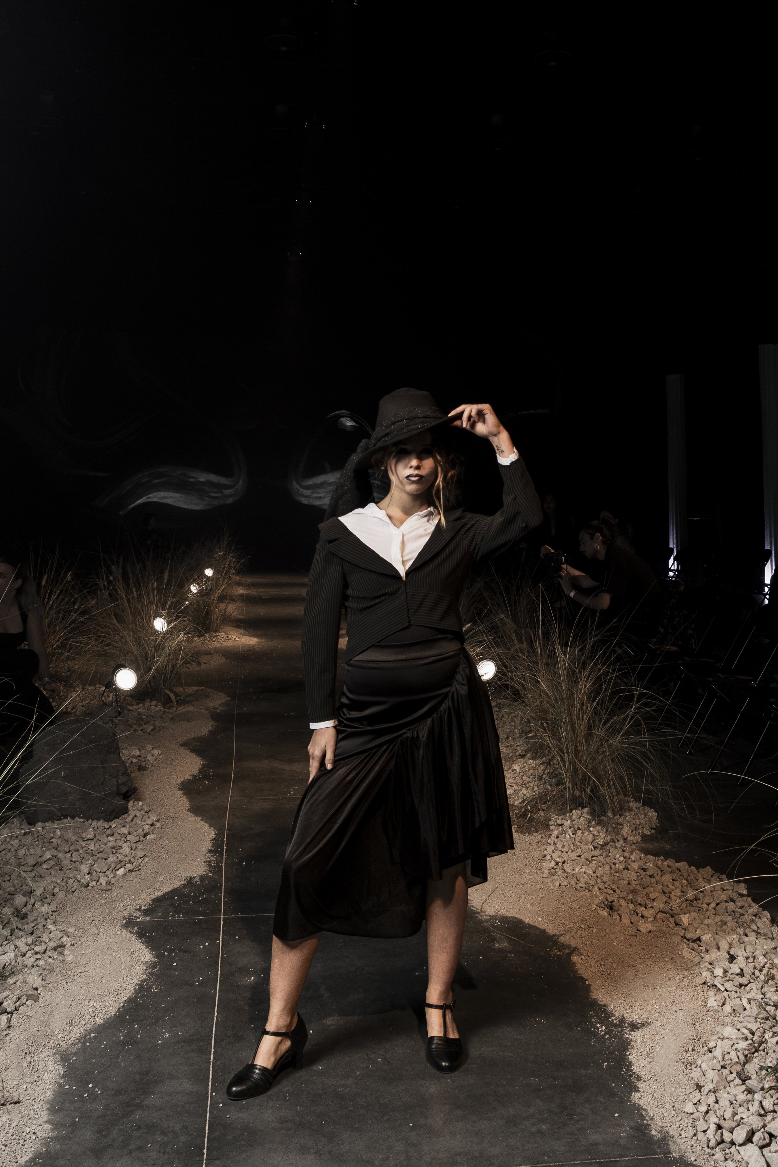 A woman in a fashionable black and white outfit standing on a runway at night, wearing a large black hat, in a desert-like setting with lights and dry grass.