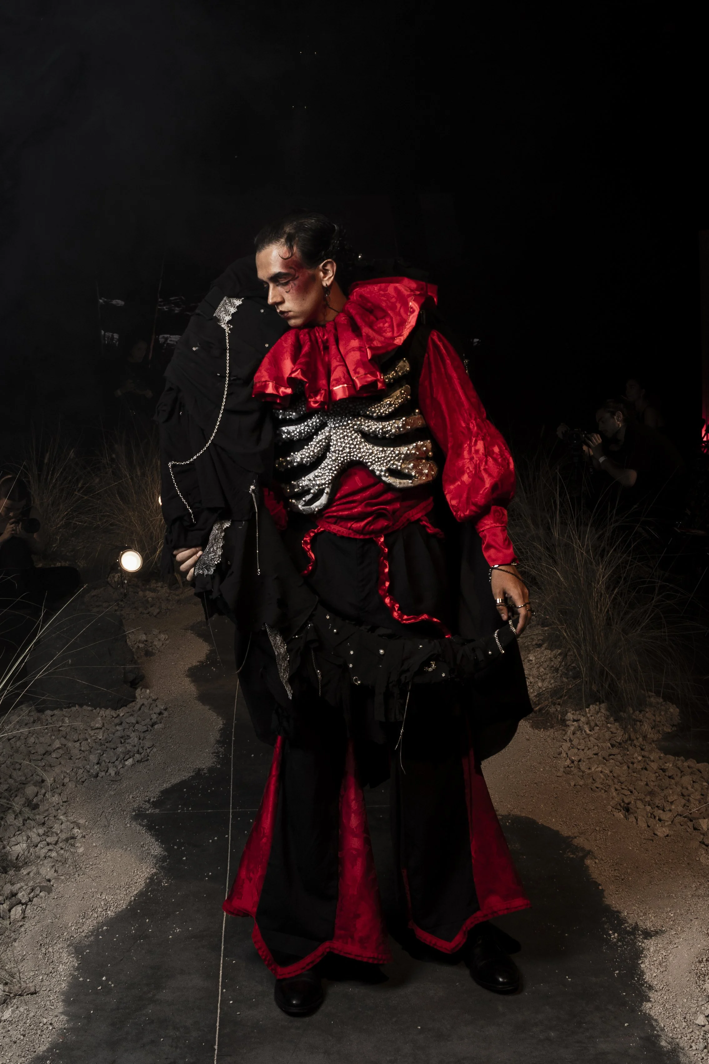 A person dressed in a dramatic black and red outfit with embellished gloves, standing outdoors at night with a dark background and some photographers in the distance.