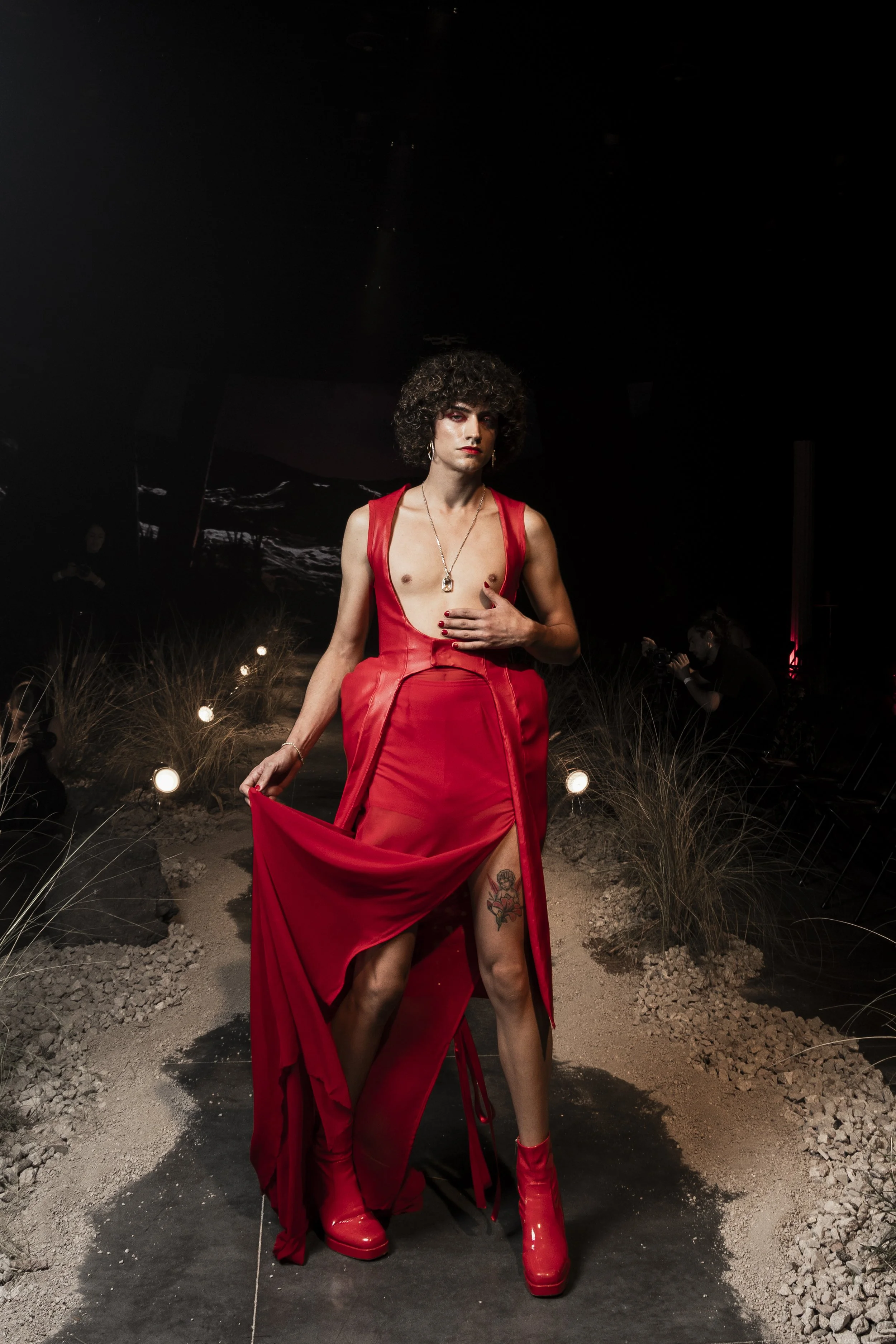 A person with curly hair wearing a red dress and boots walking on a runway with spotlights and a dark background.