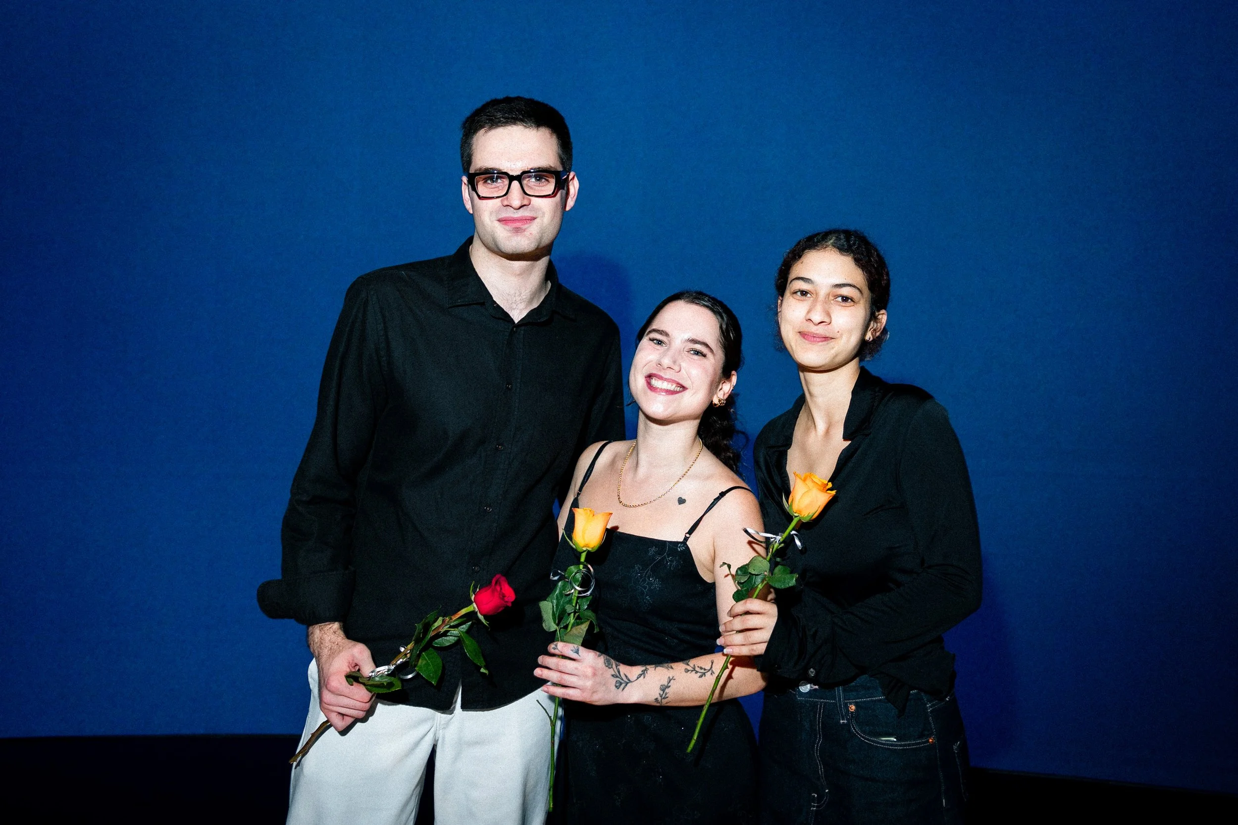 Three people standing against a blue background, smiling, holding roses, dressed in dark and light clothing.