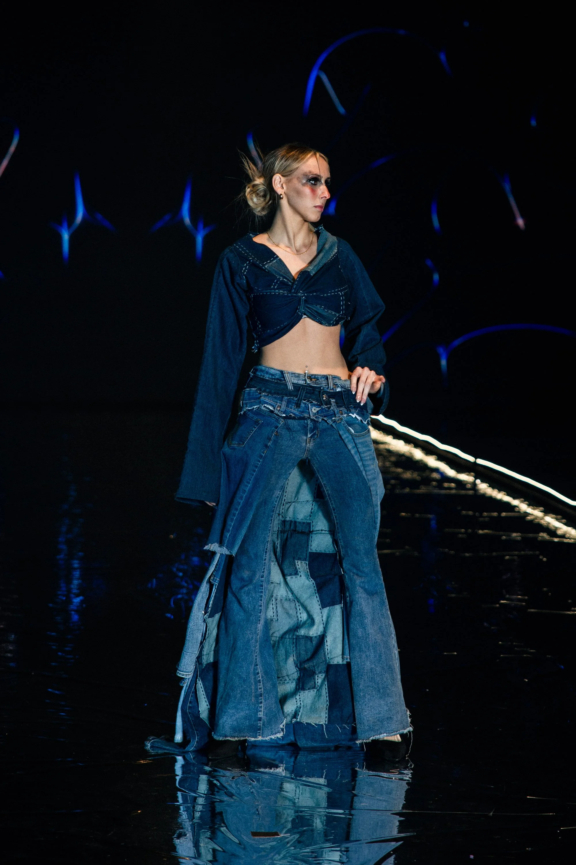 Model walking on a runway wearing a dark denim cropped jacket and patchwork denim wide-leg pants, with dark makeup and styled hair, in a dimly lit setting with blue lighting accents.