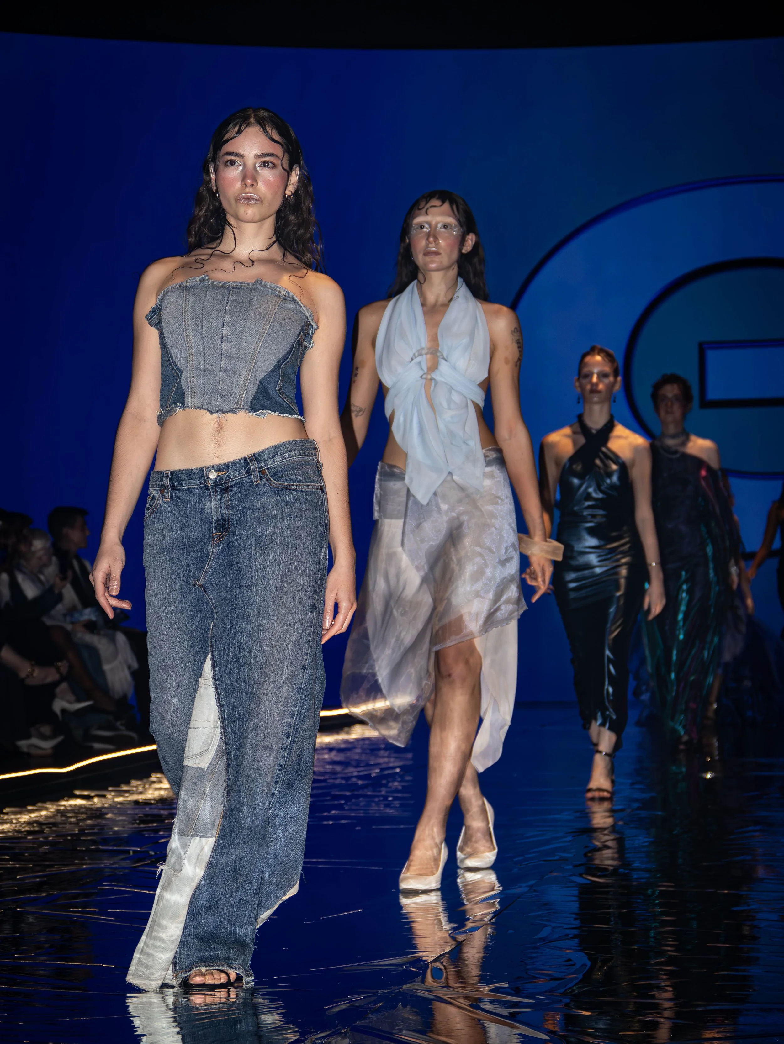 Models walking on a runway during a fashion show, dressed in various outfits, against a dark blue background with a large circular design.