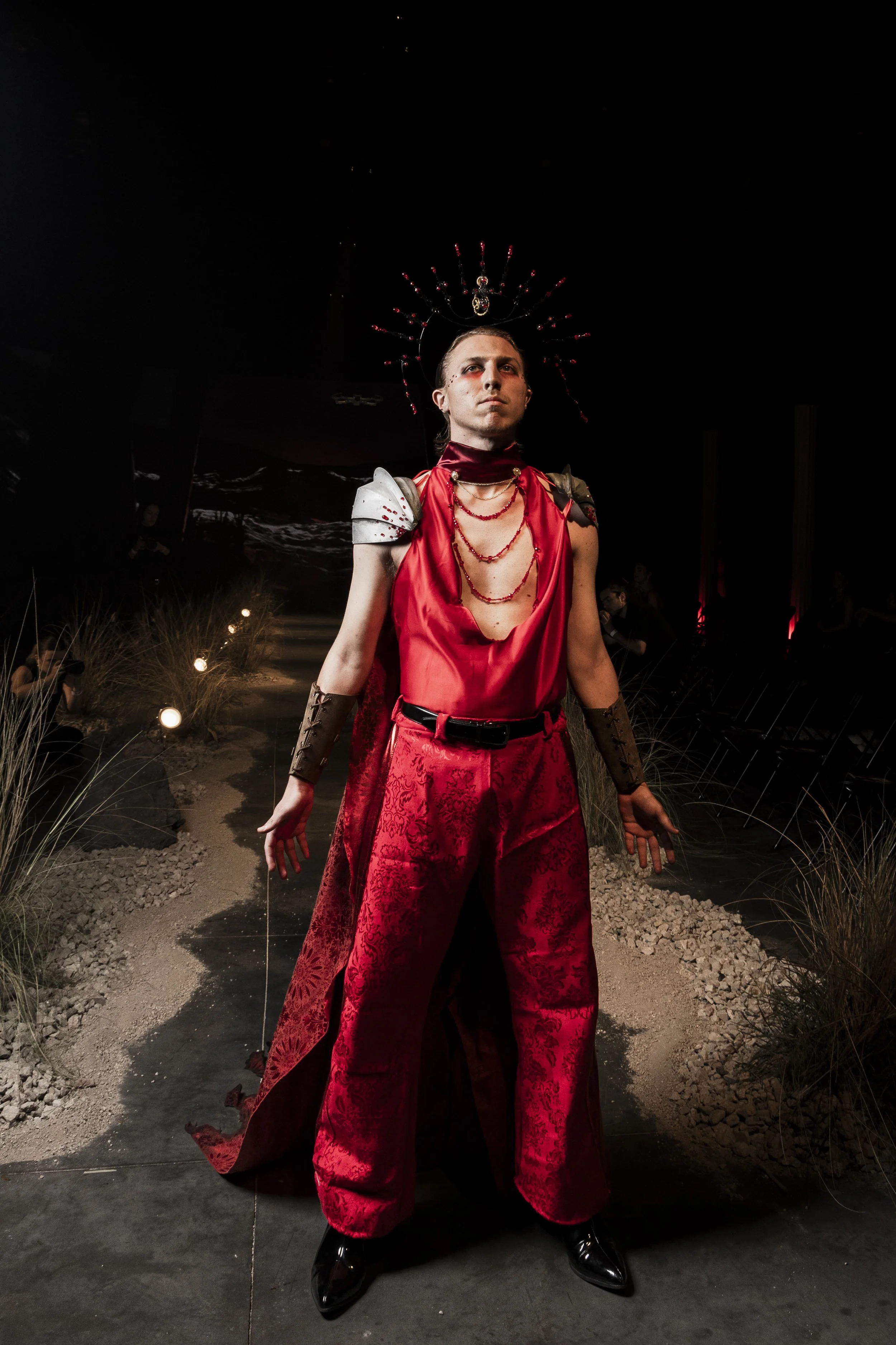 A person dressed in a dramatic red and black costume with ornate accessories and a headdress, standing on a runway at night.