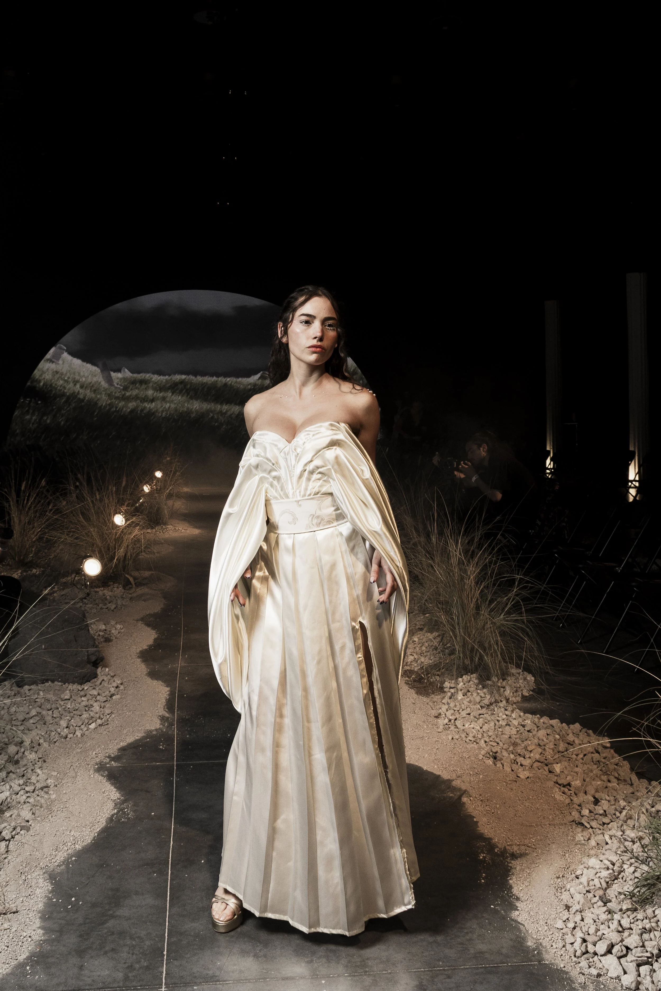 A woman wearing an elegant, strapless, cream-colored gown with puffy sleeves stands on a dark runway with a scenic backdrop of a cloudy sky and grassy landscape. She has dark hair and a serious expression. There is a photographer in the background.