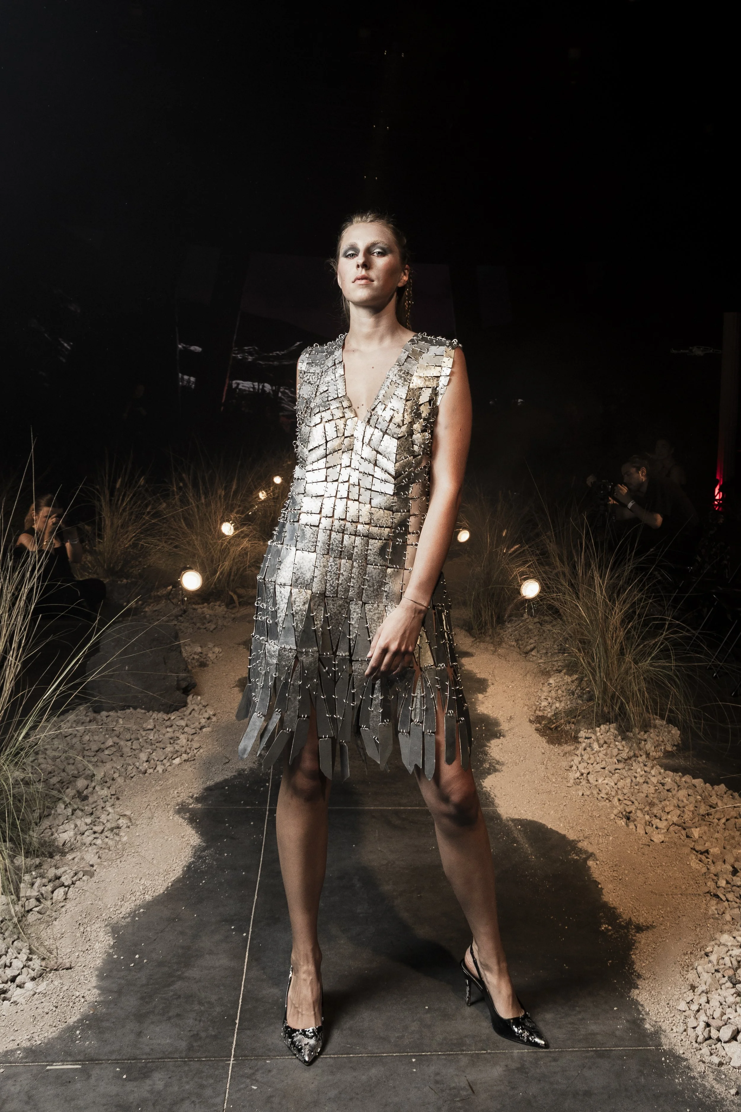 A woman wearing a metallic, reflective dress made of rectangular metal plates standing in a dark outdoor setting with grass and small lights, with photographers and spectators in the background.