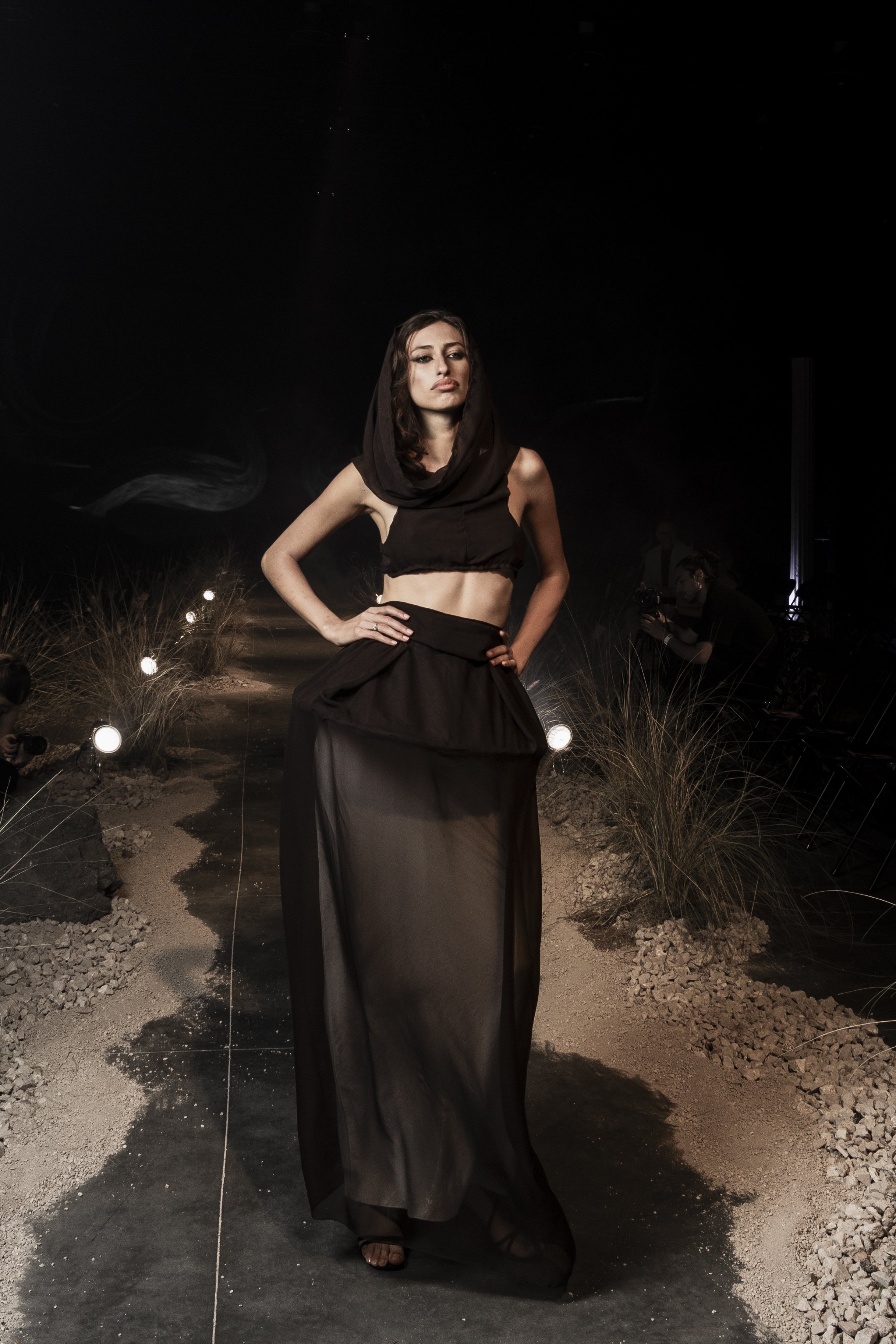 A woman with dark wavy hair wearing a black sleeveless crop top and a long black sheer skirt, standing outdoors at night on a pathway surrounded by dry grass and small rocks, with lights and people visible in the background.
