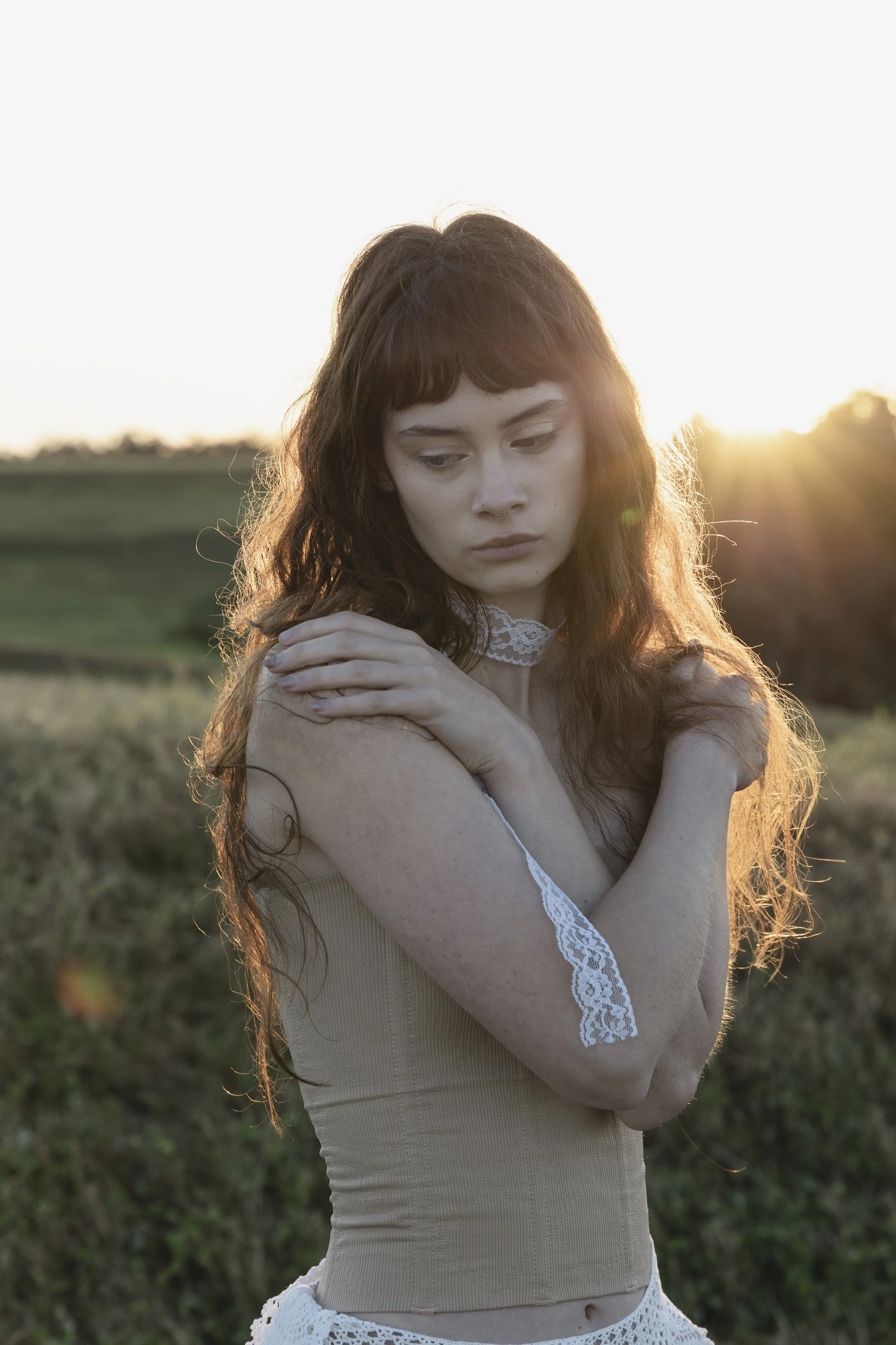 A young woman with long, wavy brown hair stands outdoors during sunset, looking down with a contemplative expression. She wears a beige sleeveless top with lace details on the shoulders and a choker. Her arms are crossed over her chest, and the sun is setting behind her, casting a warm glow.