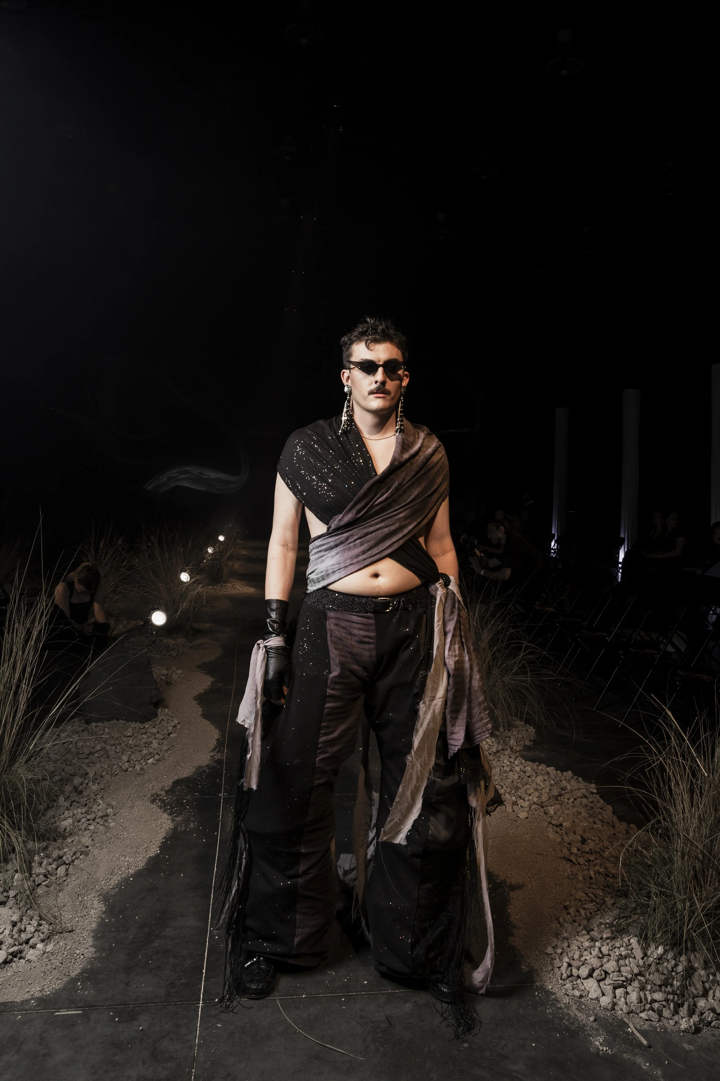 A person with short dark hair, wearing sunglasses, large earrings, and a black sparkly top draped with a brown fabric, standing on a pathway surrounded by tall grass and rocks, at night with a dark sky and minimal background lighting.