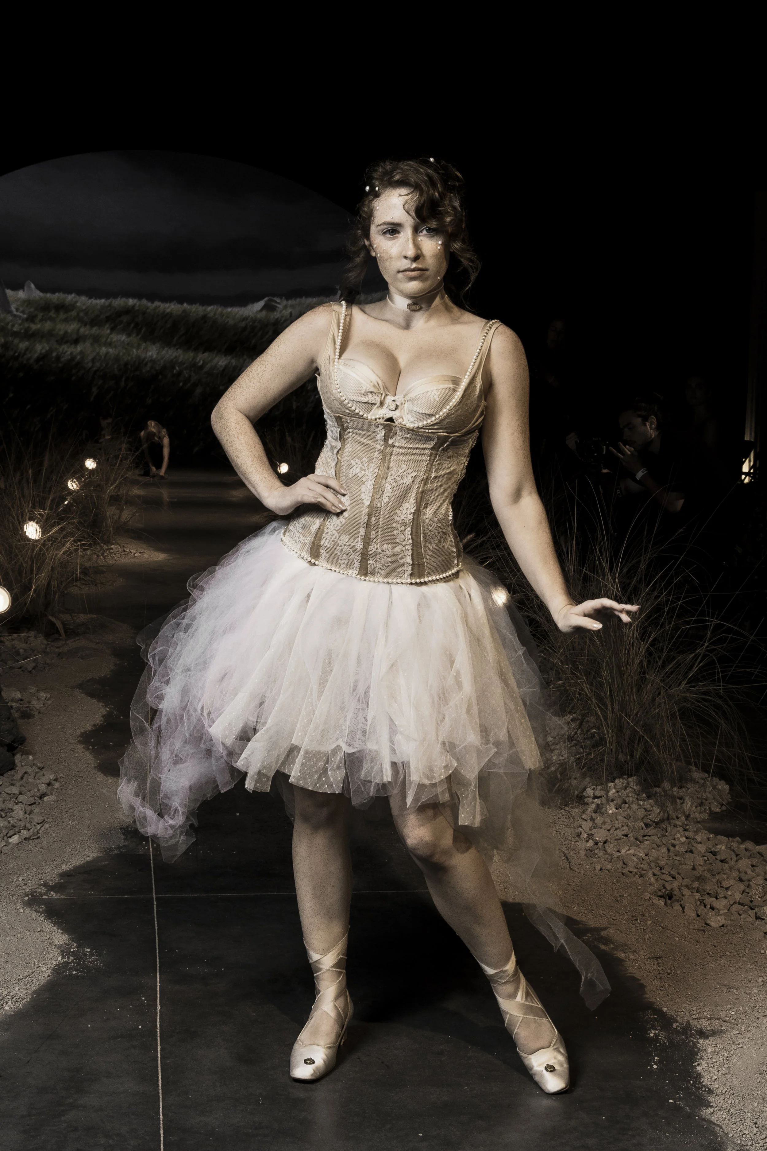 A person dressed as a ballerina standing on a black stage with a dark background that shows a landscape of tall grass and a cloudy sky.