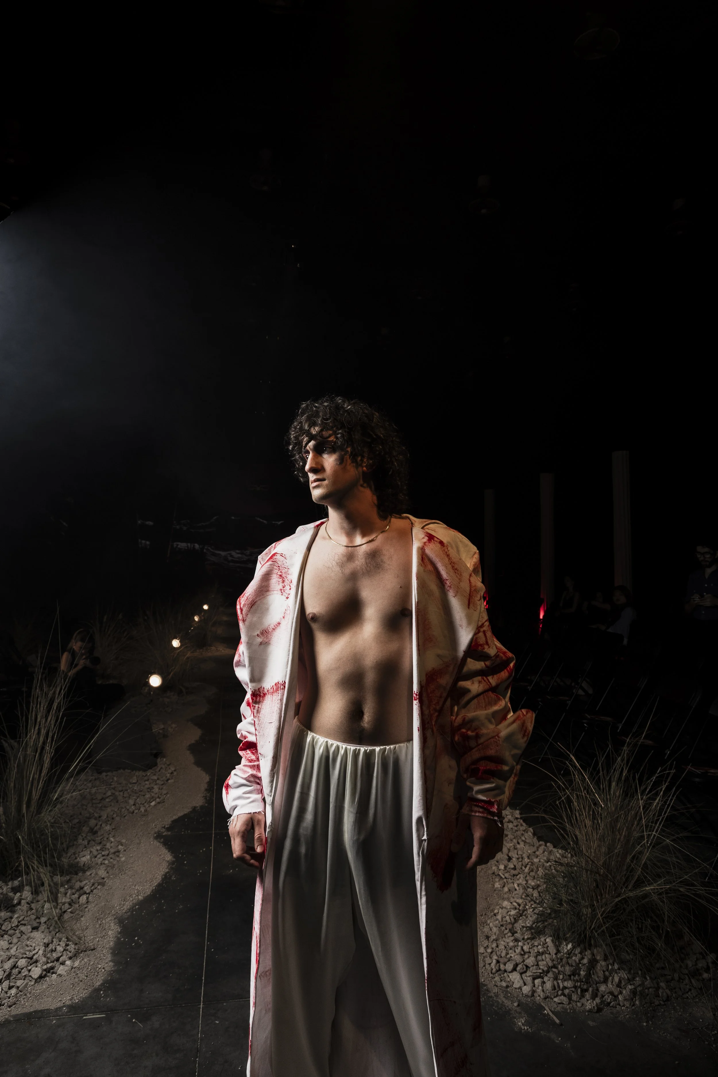 A man with curly hair, wearing open silk pants and a blood-stained satin robe, standing outdoors at night under a spotlight.