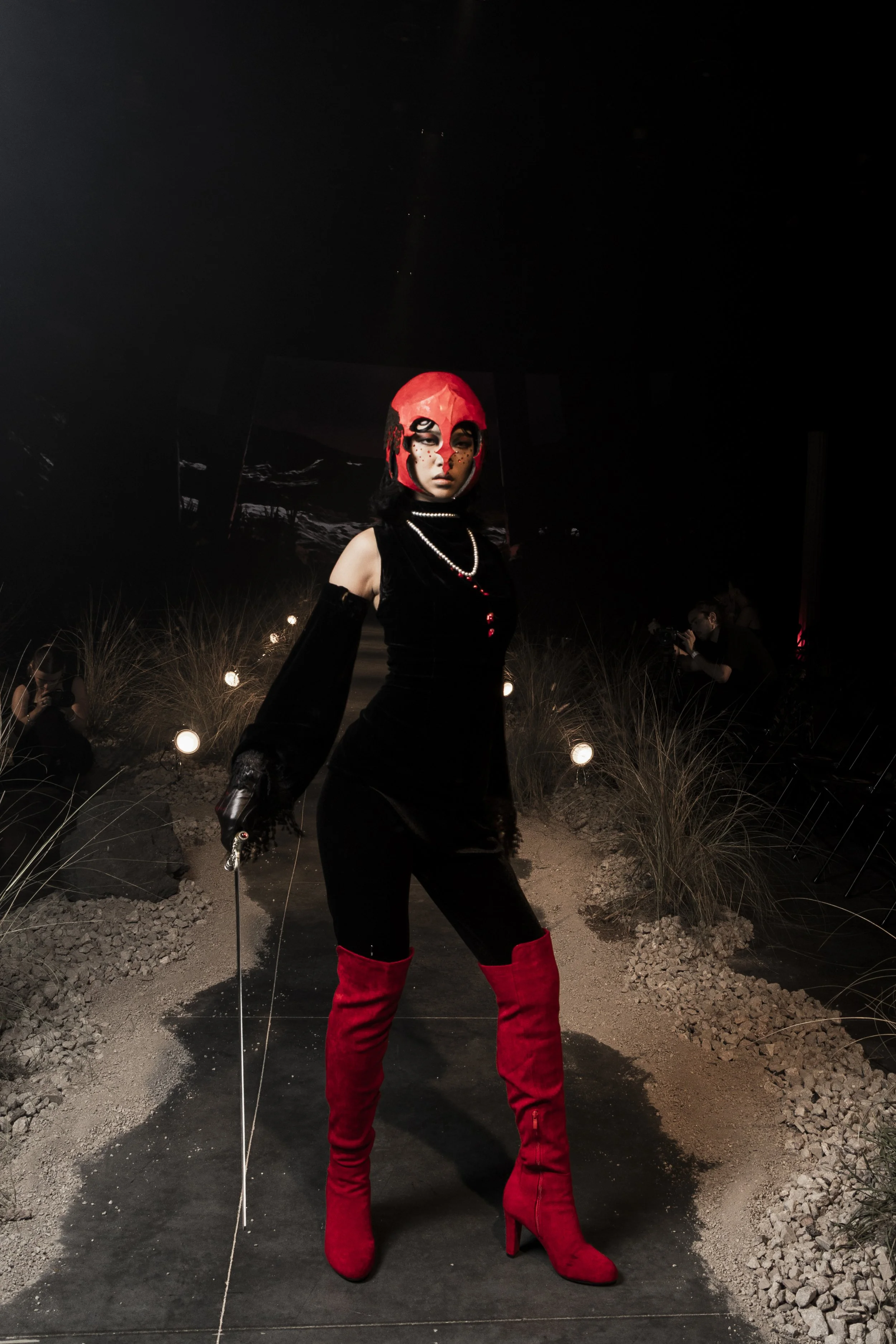 A woman wearing a red helmet with black and white makeup, black sleeveless top with pearl necklaces, black gloves, red boots, and black pants standing in a dimly lit outdoor setting with small lights and plants.