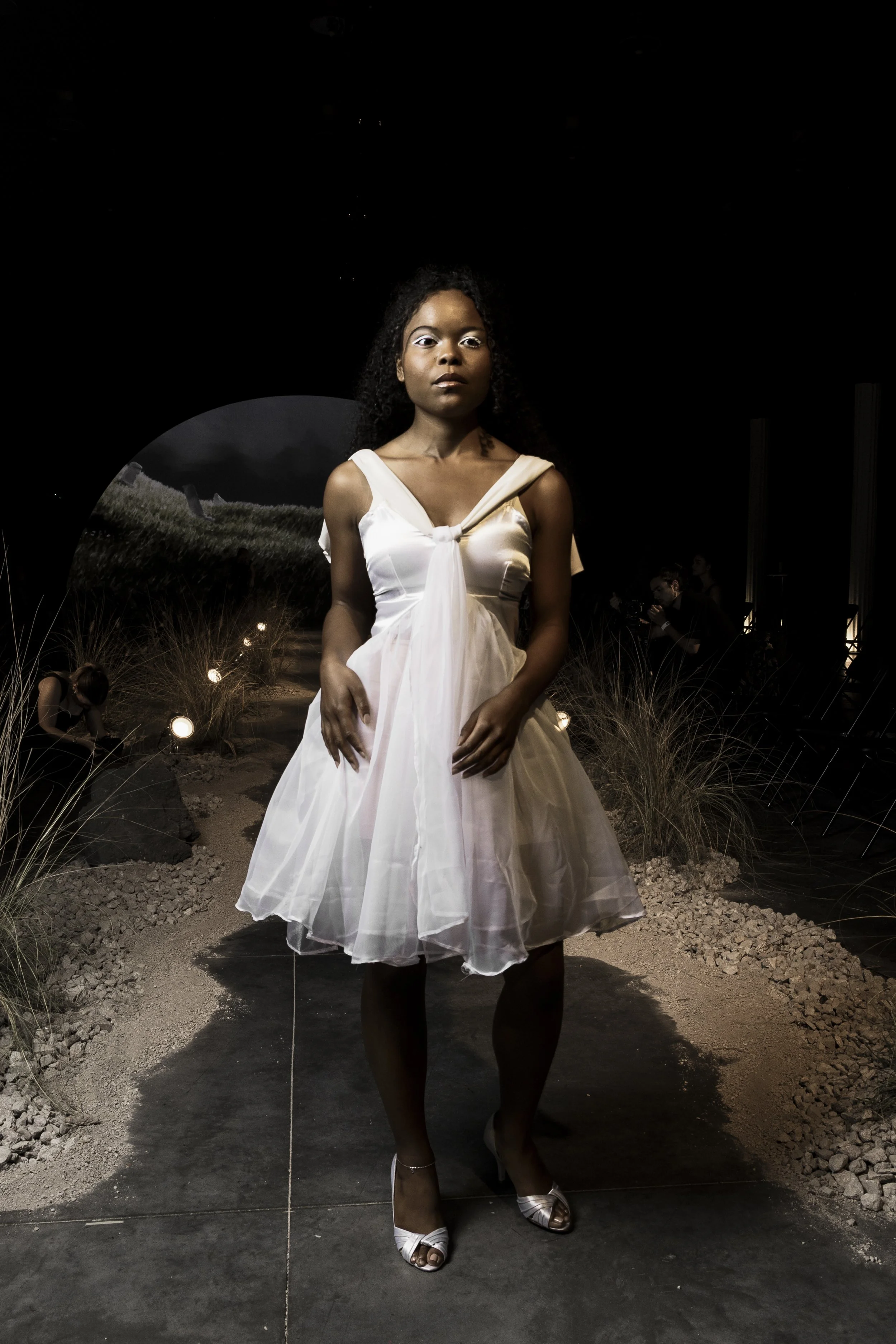 A woman in a white, elegant dress with a full skirt and tied front, standing on a dark runway with spotlights and landscape scenery in the background, at an indoor fashion event.