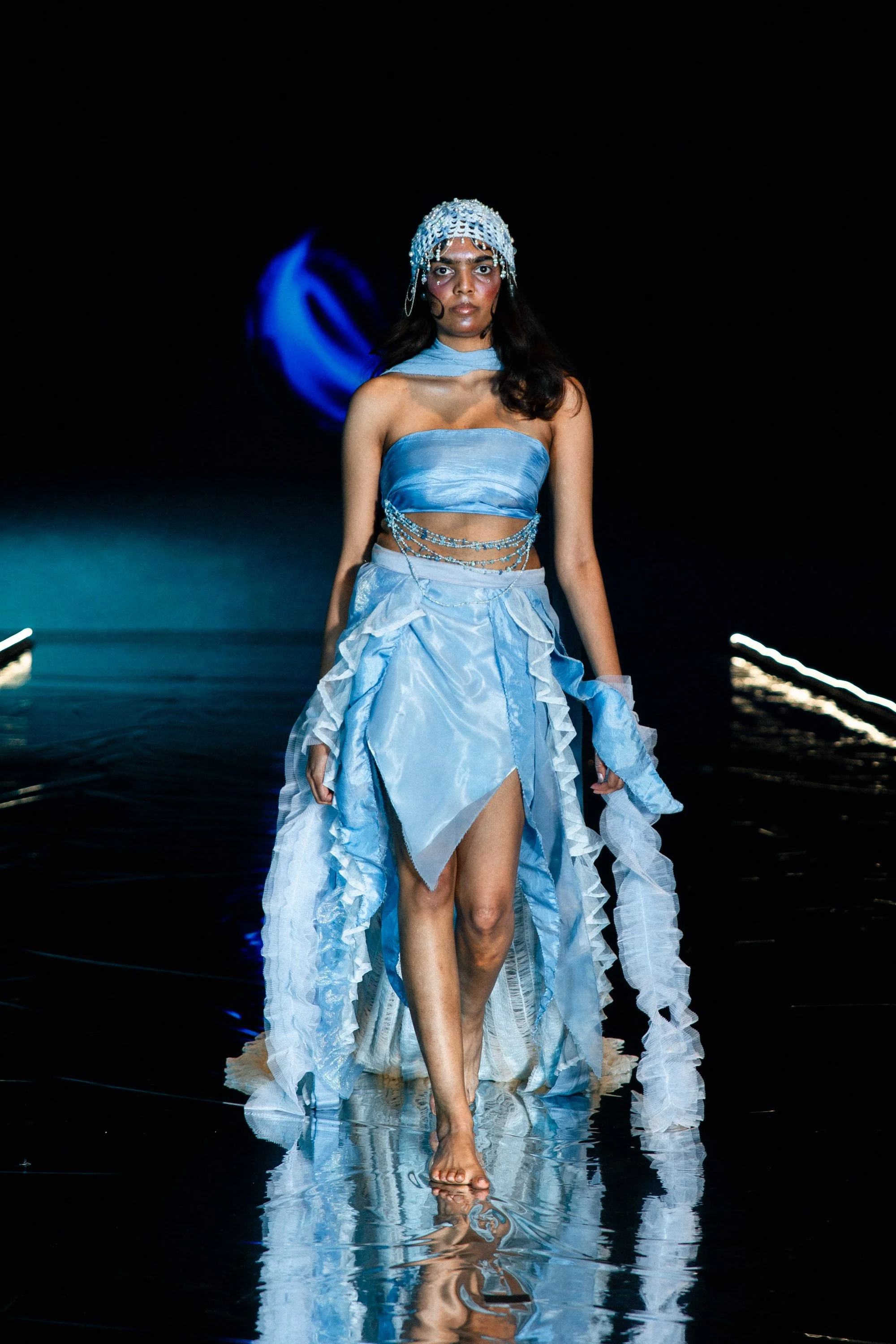 A model walking barefoot on a runway in a light blue, ruffled, strapless dress with chain embellishments, wearing a matching headpiece with beads and sequins, against a dark background with reflections on the runway.