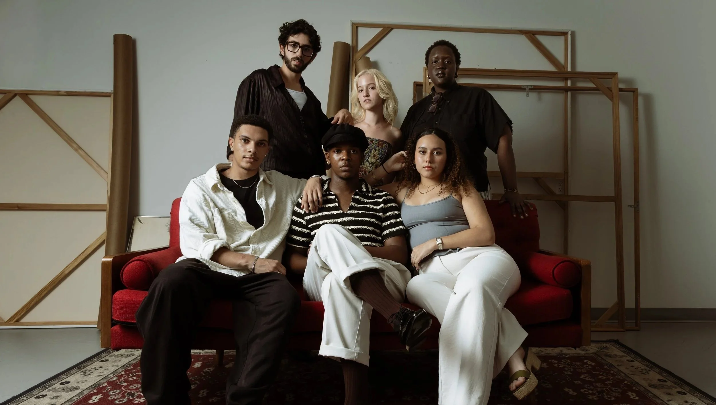 Group portrait of six diverse young adults sitting and standing on and around a red sofa in a room with plain walls and wooden frames.