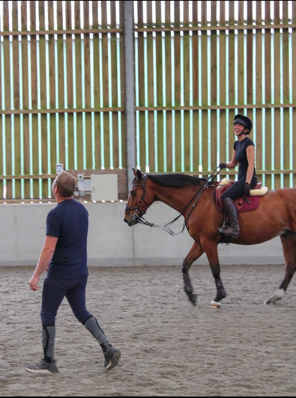 A woman riding a horse while smiling and wearing a helmet inside an indoor riding arena, with a person walking nearby.