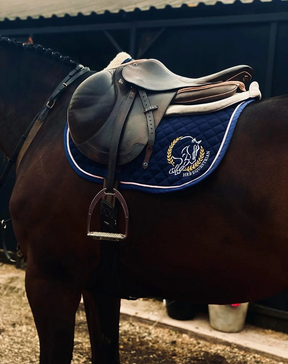 A close-up of a dark horse with a saddle, bridle, and blue saddle pad with a logo and the text 'HRB Equstrian' in gold, in a stable or riding arena.