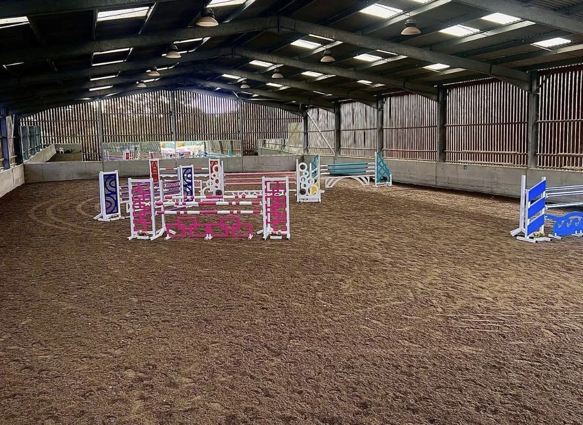 Indoor horse riding arena with colorful show jumping obstacles set up on a dirt floor, covered by a metal roof with skylights.