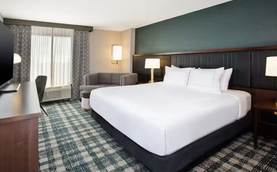 Hotel room with a large bed with white linens, two bedside lamps, a gray armchair, a window with curtains, a desk with a chair, and a patterned carpet.